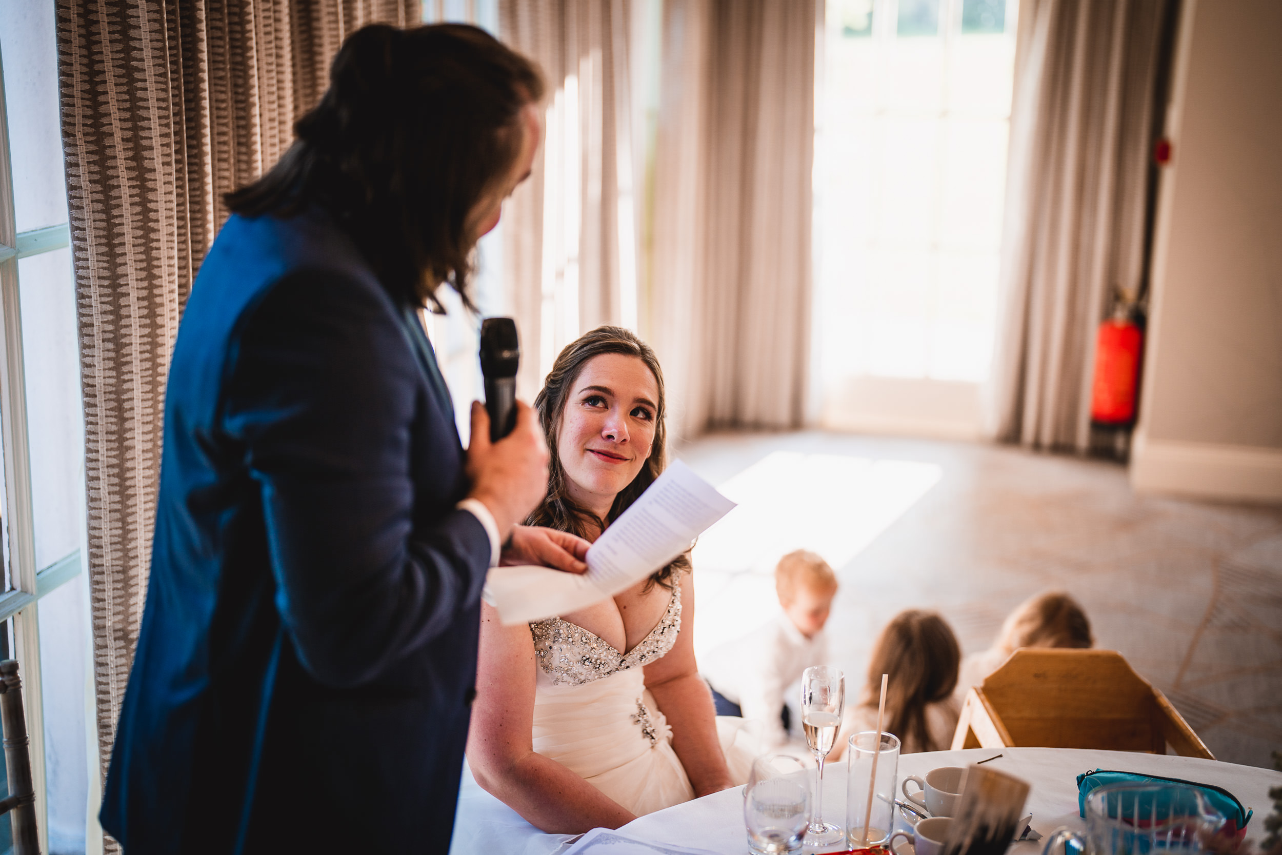 A person in a blue suit speaks into a microphone, holding a piece of paper. A woman in a white dress sits, listening attentively at a table with dishes and glasses.