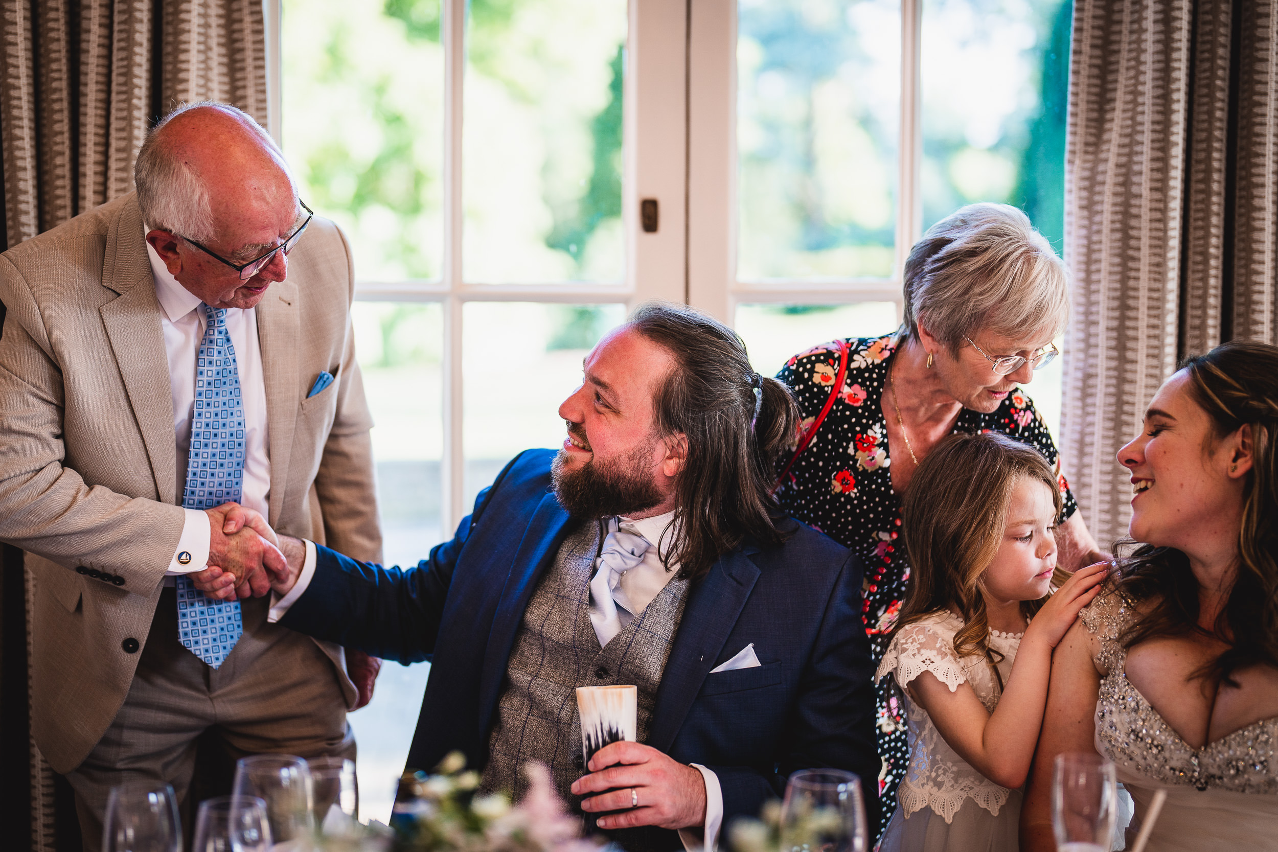 A man in a suit shakes hands with another man at a table, while a woman stands nearby with a young girl. Another seated woman looks on, holding a child.