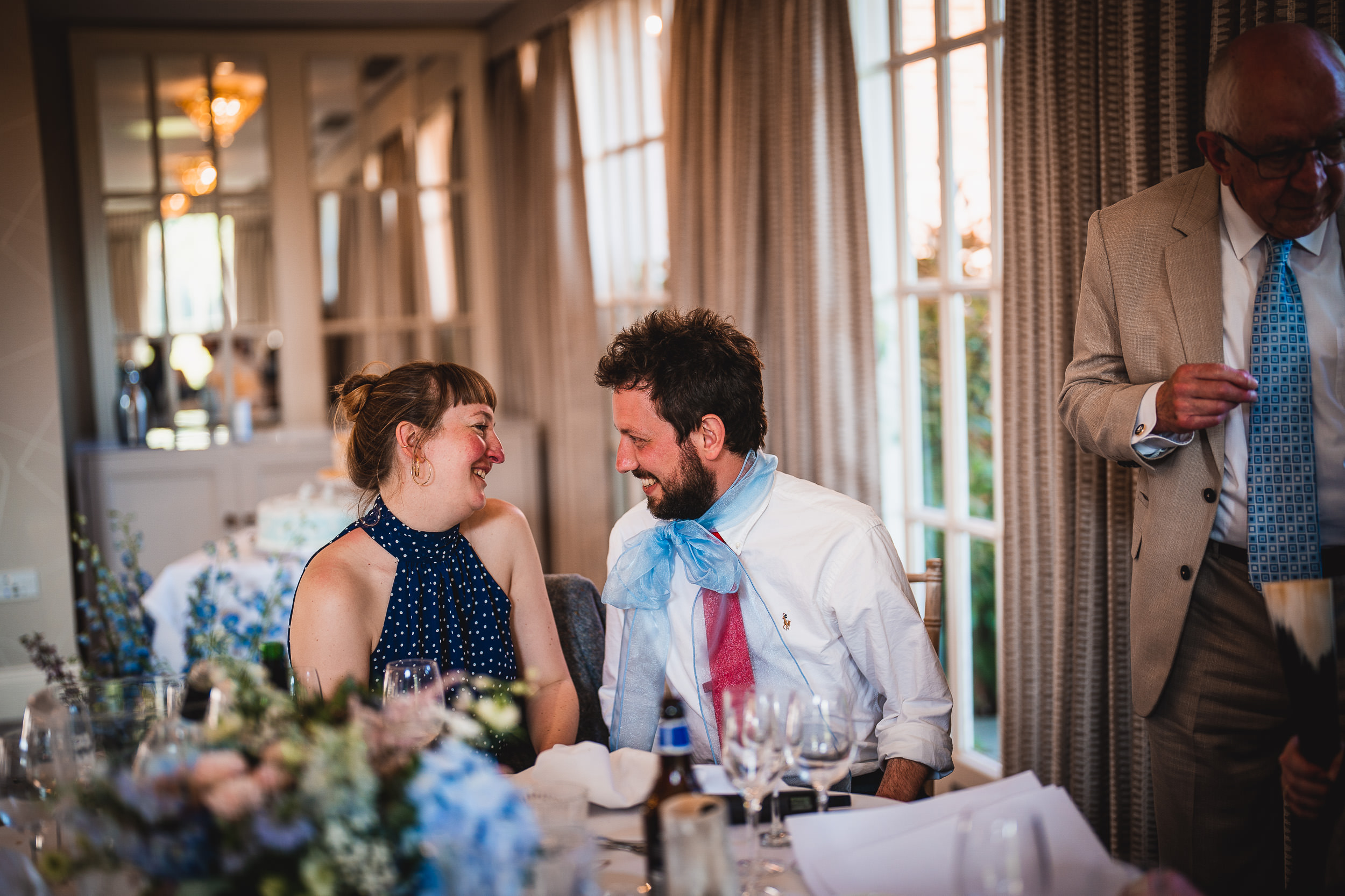 A smiling couple sits at a dining table covered with glasses and flowers, gazing at each other. An older man stands nearby, adjusting his jacket.