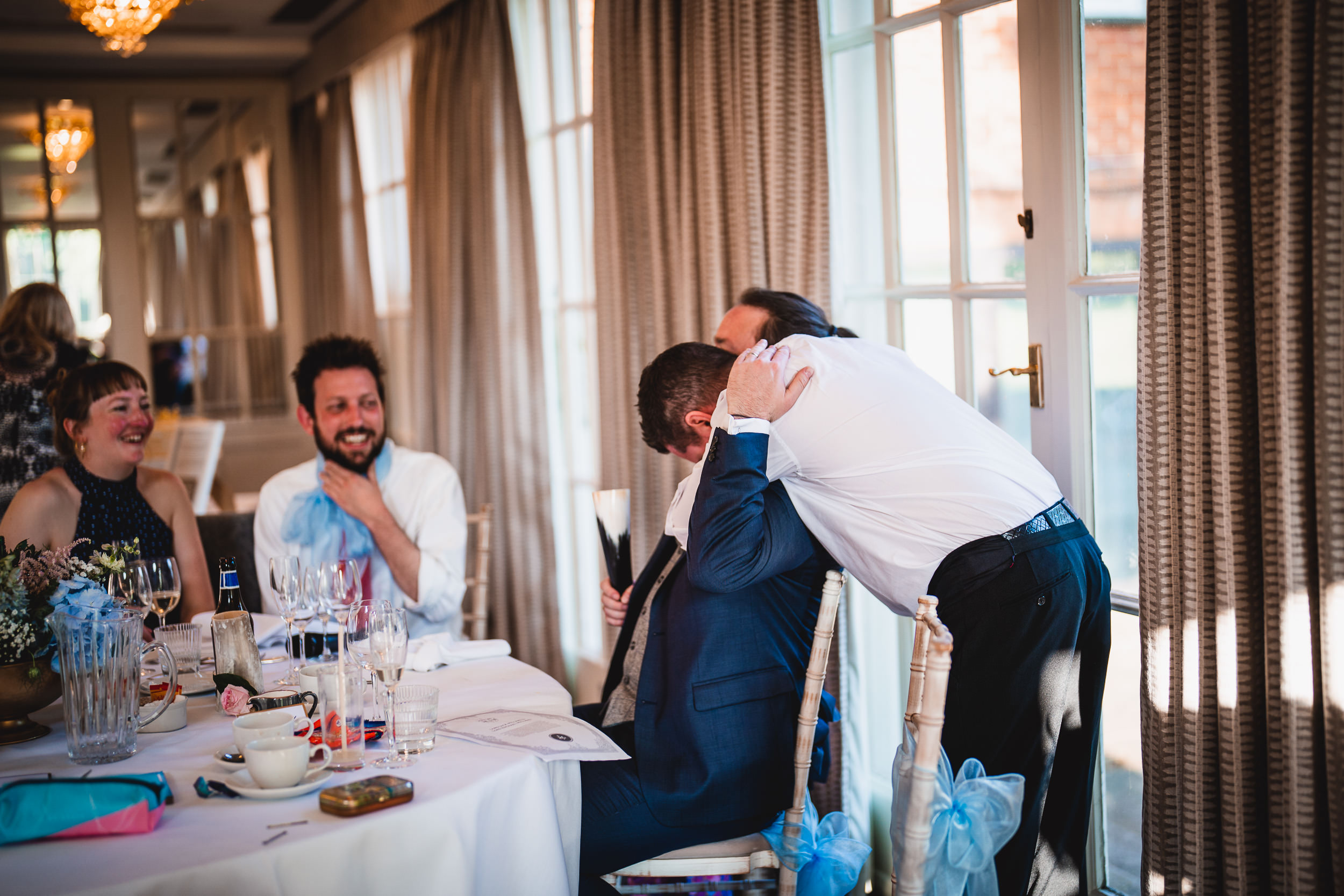 A man in a white shirt leans over another man in a blue suit at a dining table, with guests laughing and curtains in the background.