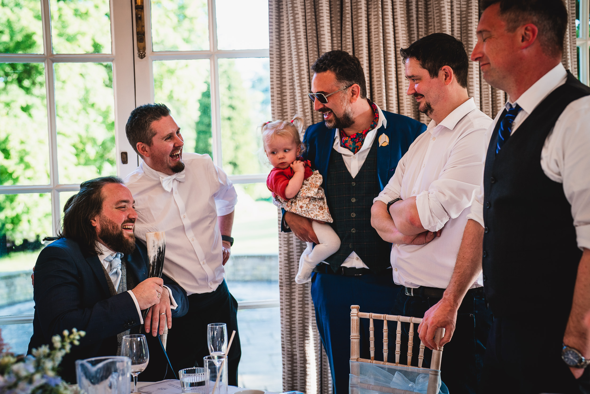 A group of men stand around a table, smiling and talking. One man is seated, holding a glass, while another holds a small child in red. Bright window and curtain backdrop.