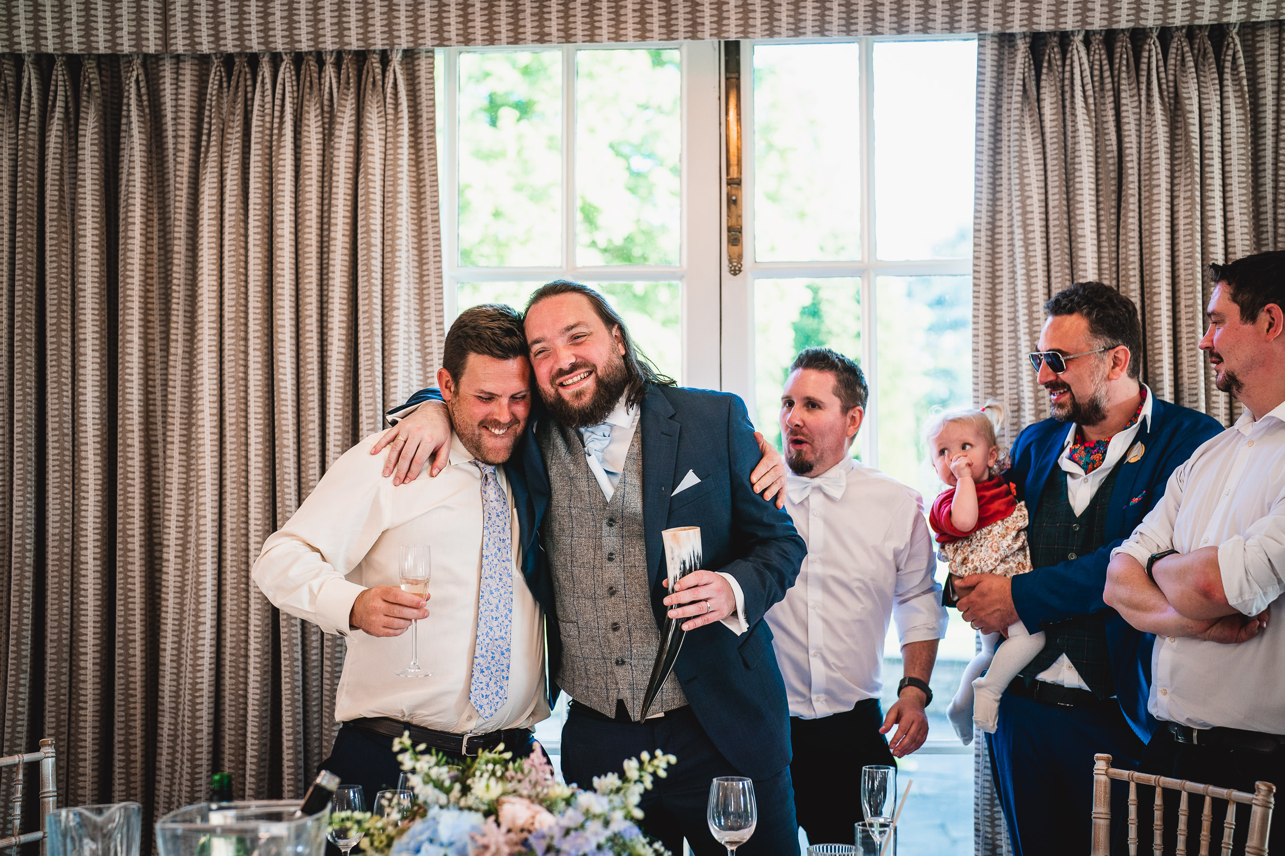 A group of five men, one holding a baby, stand together smiling in a room with a large window and curtains. They appear to be celebrating, holding drinks and dressed in formal attire.