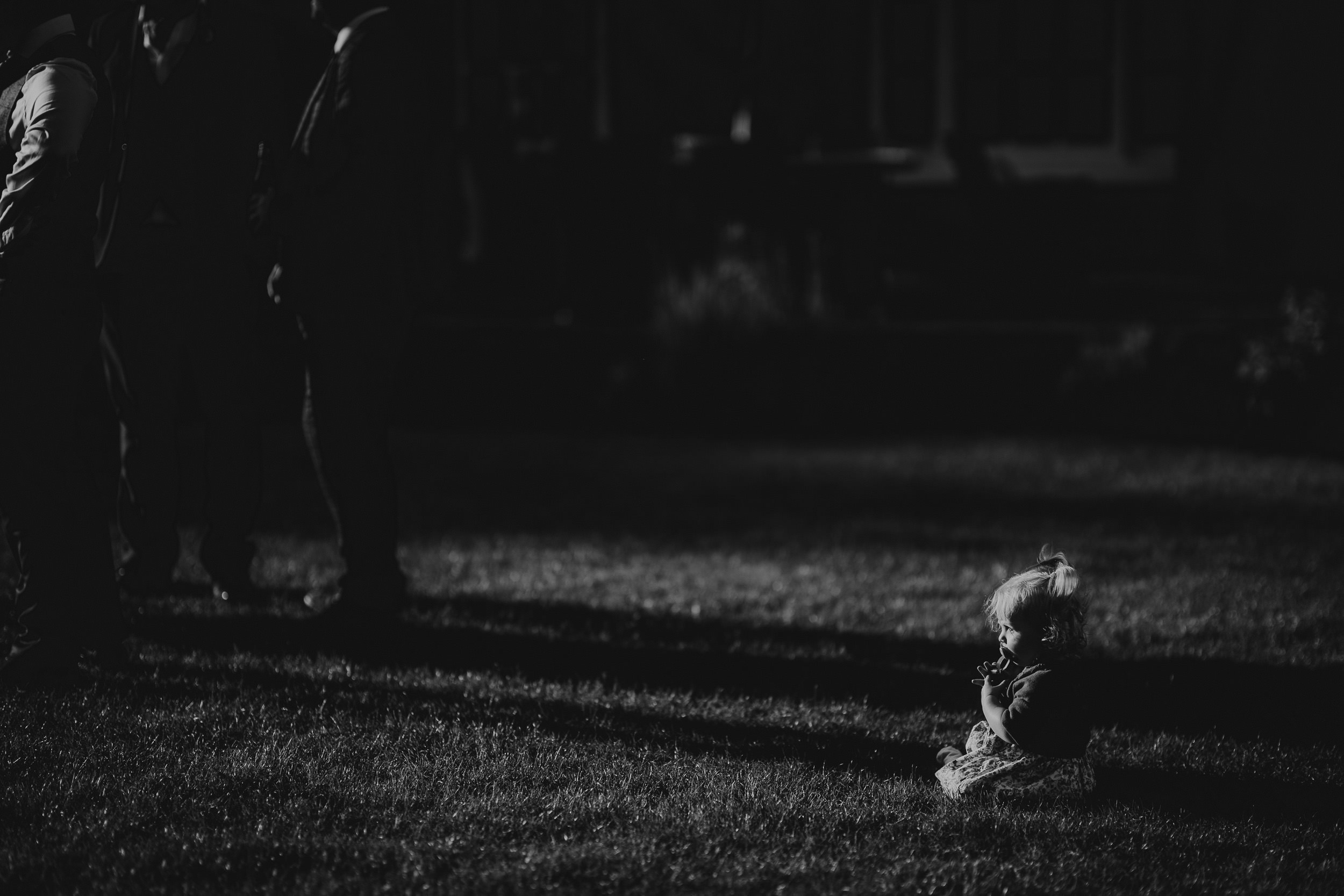 A small child sits alone on a grassy field at night, surrounded by shadows of standing adults.