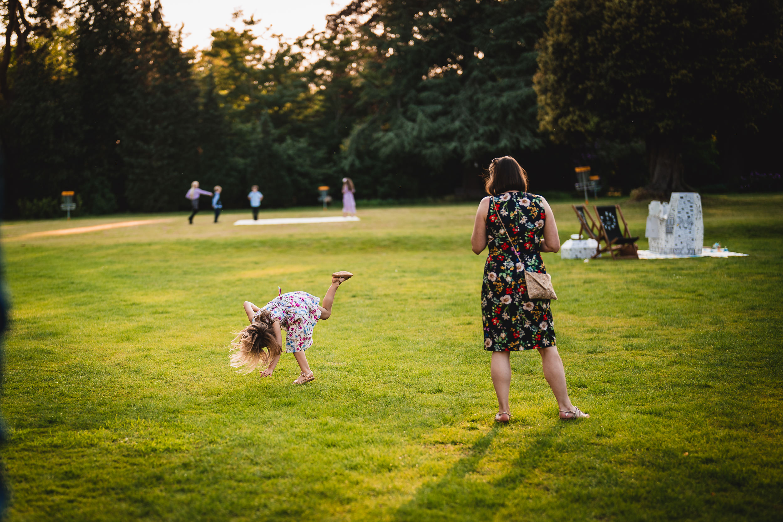 A child cartwheels on a grassy field while an adult watches. Other people are in the background.