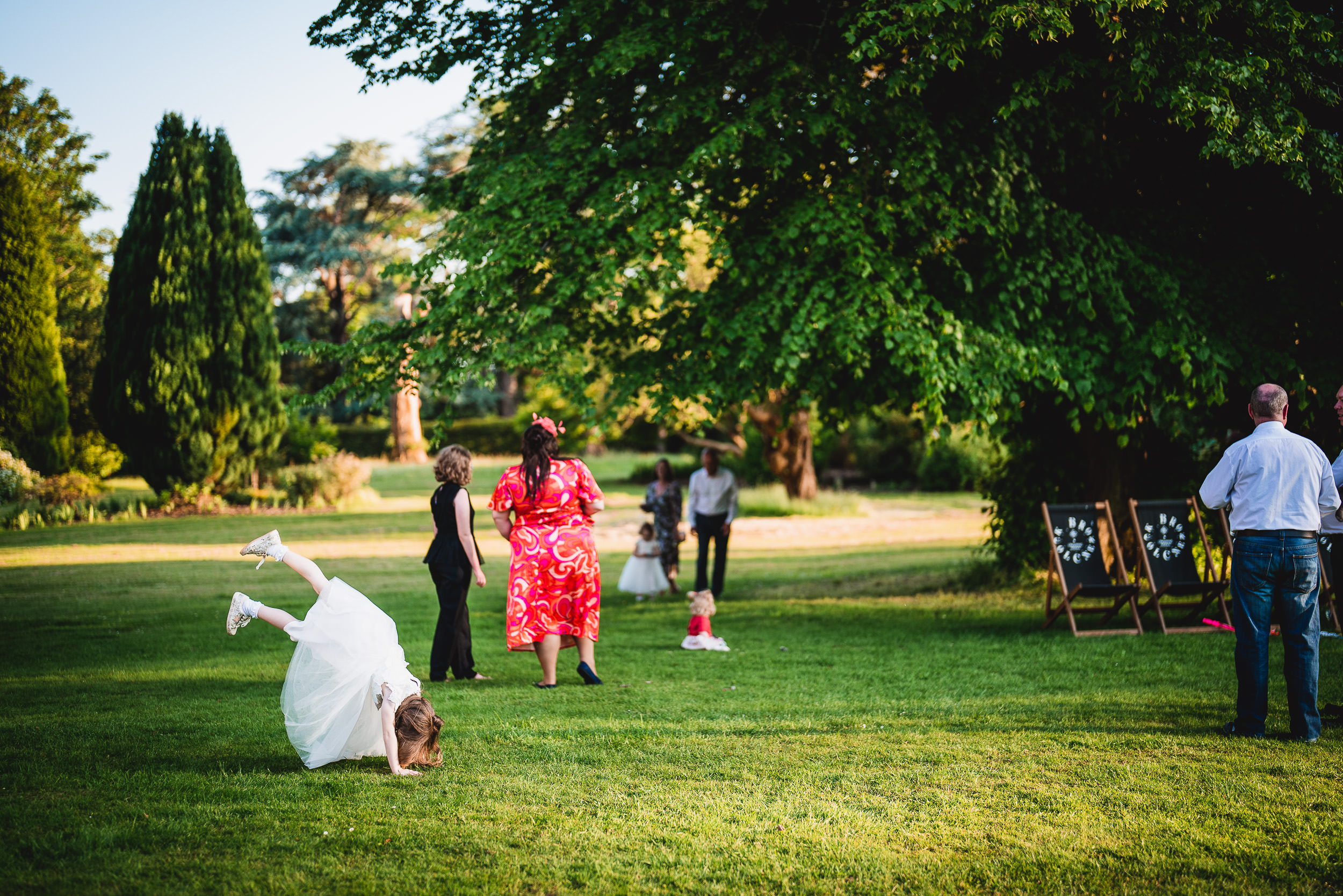 Child in a white dress doing a cartwheel on grass. Other people stand nearby under a large tree.