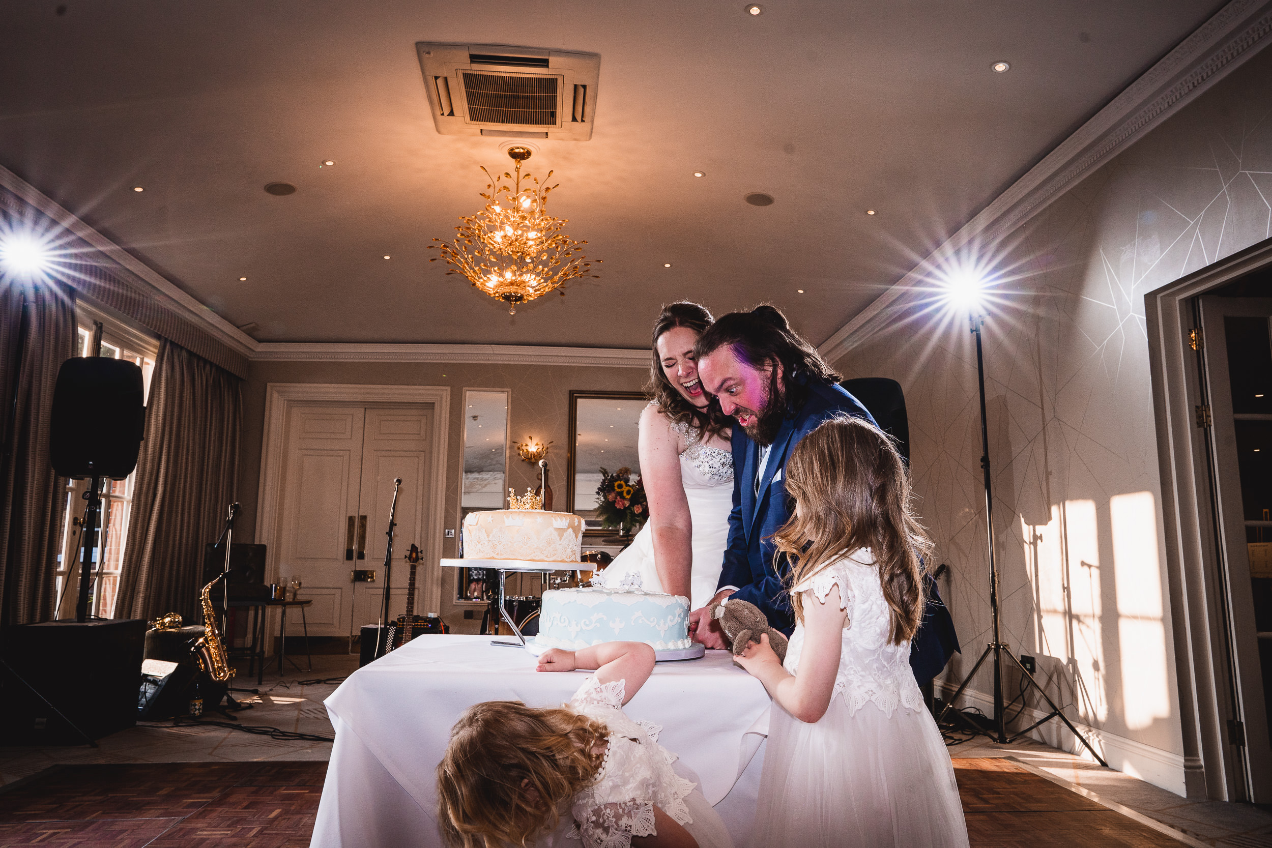 A bride and groom, along with two children, are cutting a wedding cake on a decorated table in an elegant room with chandeliers.