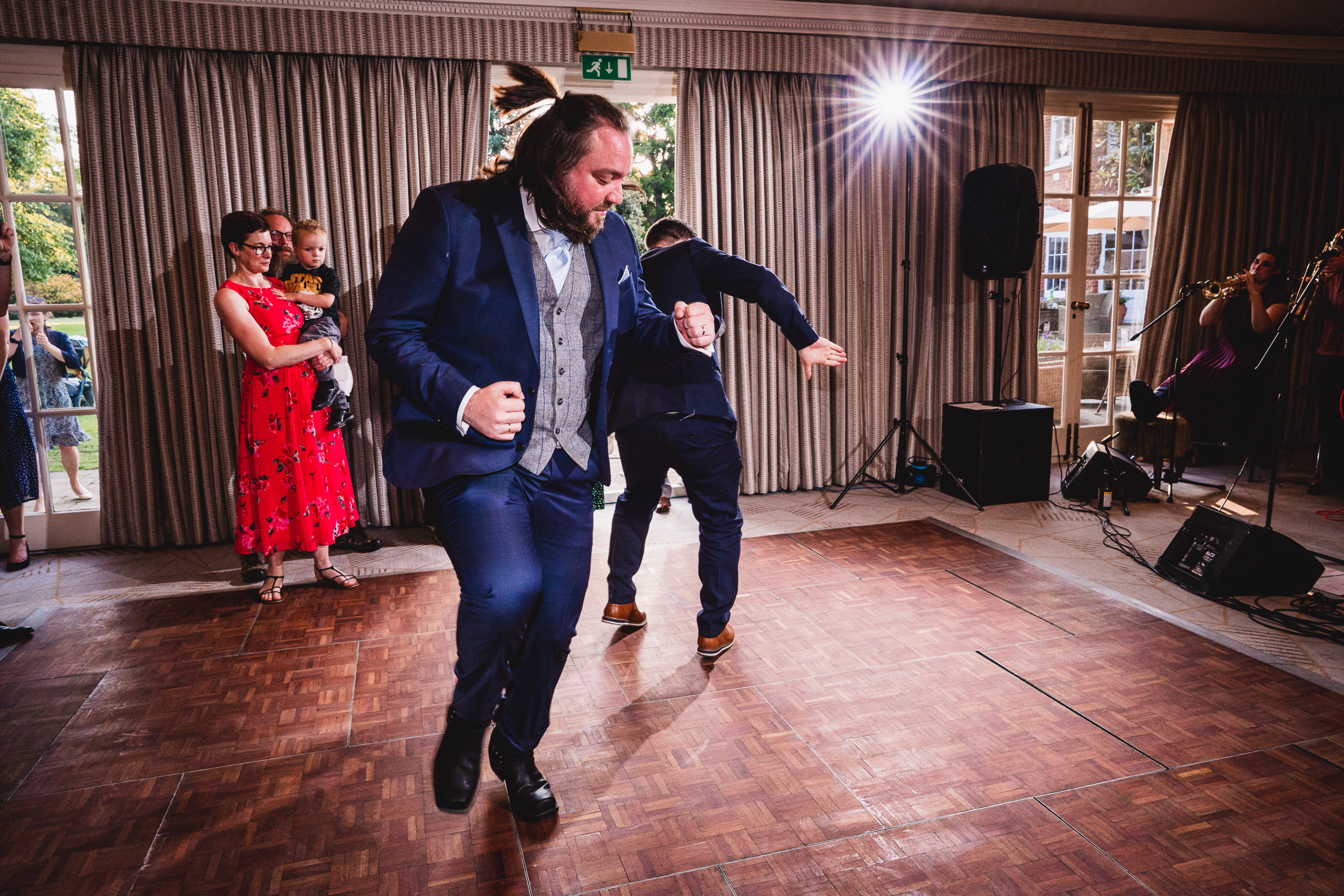 Two men in suits dance energetically on a wooden floor. Onlookers, including a woman in a red dress holding a child, watch from the side.