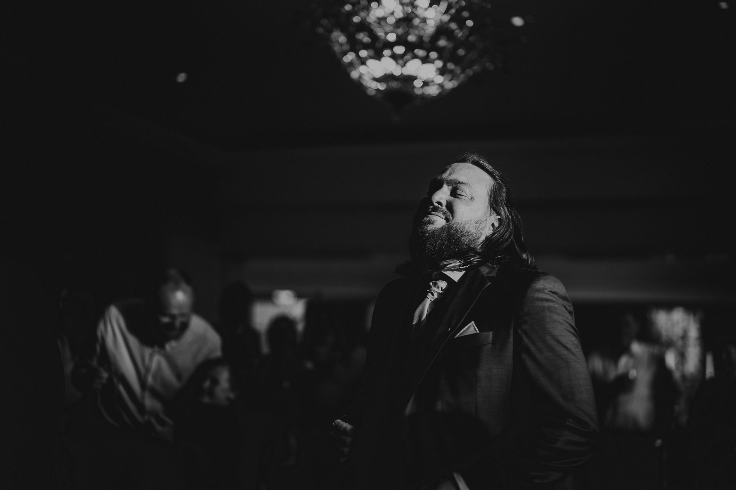 A man in a suit passionately sings under a chandelier in a dim room.