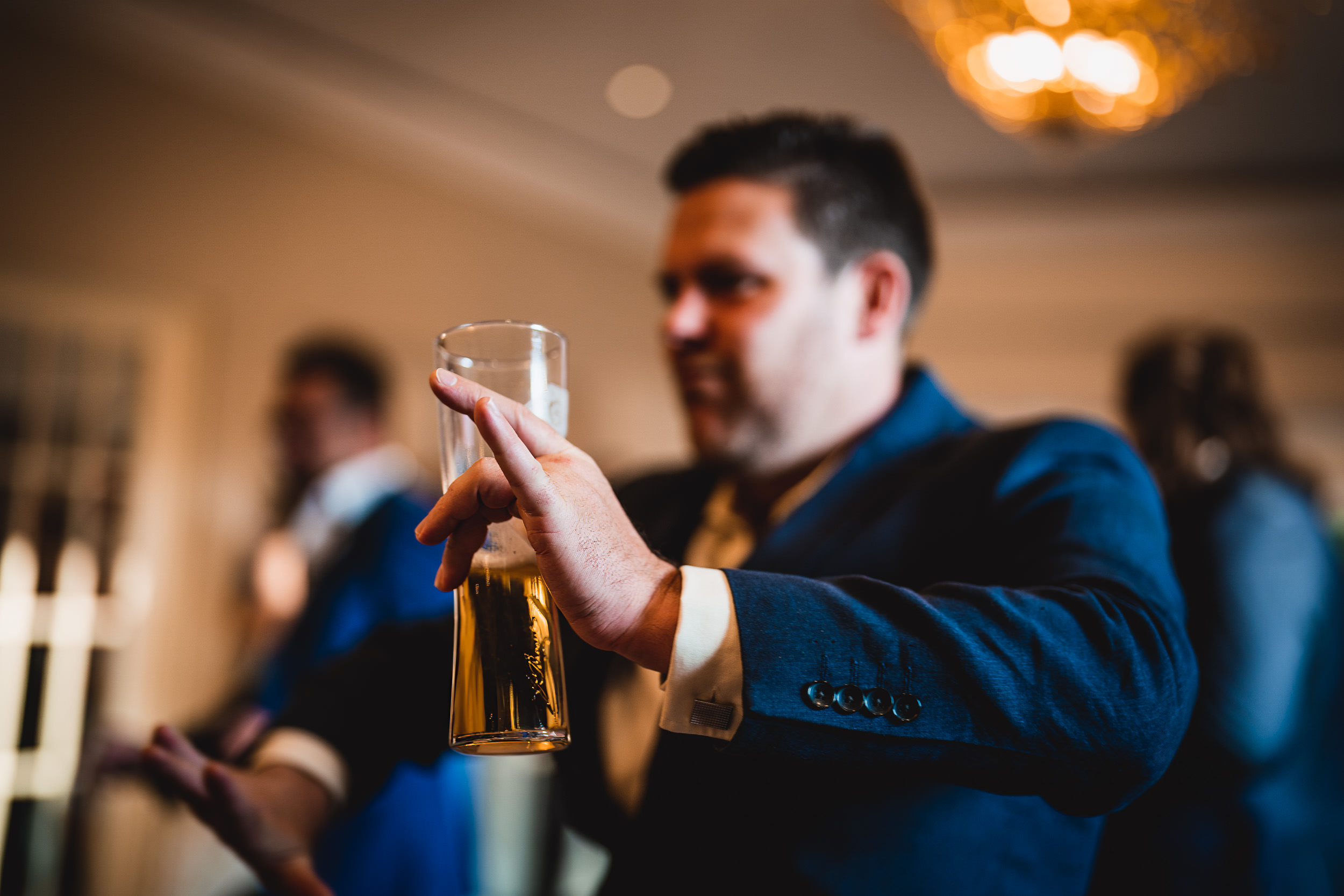 A person in a suit holds a tall glass of beer, gesturing animatedly, with blurry figures and a chandelier in the background.