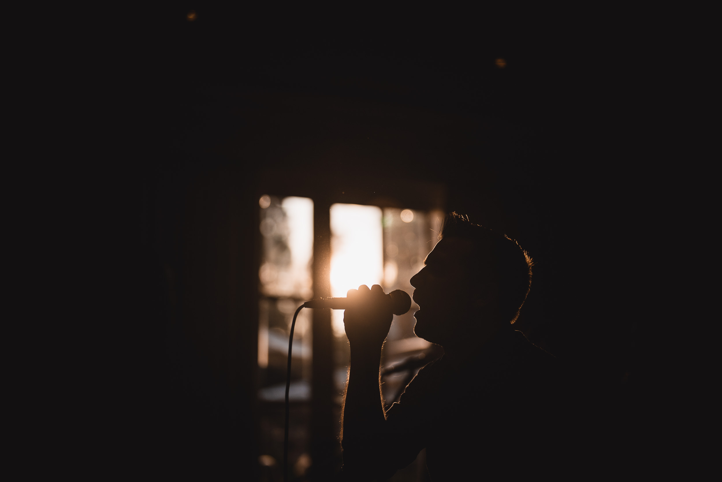 Silhouette of a person singing into a microphone with bright sunlight streaming through a window in the background.