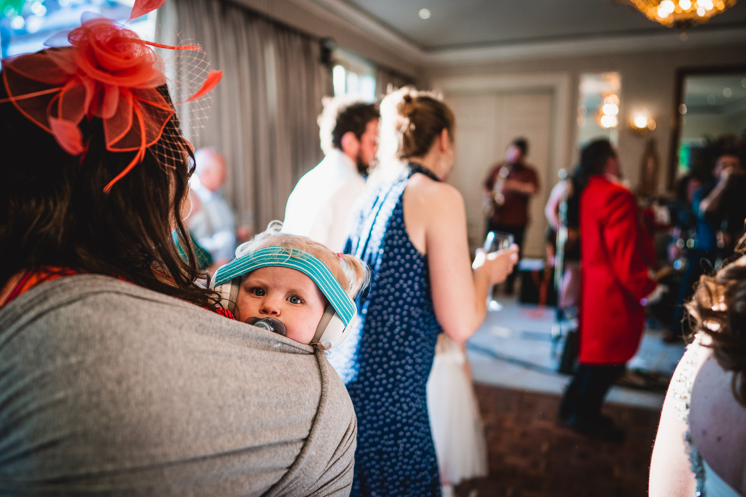 A baby wearing headphones looks over the shoulder of a woman in a lively indoor party setting with people dancing and talking.