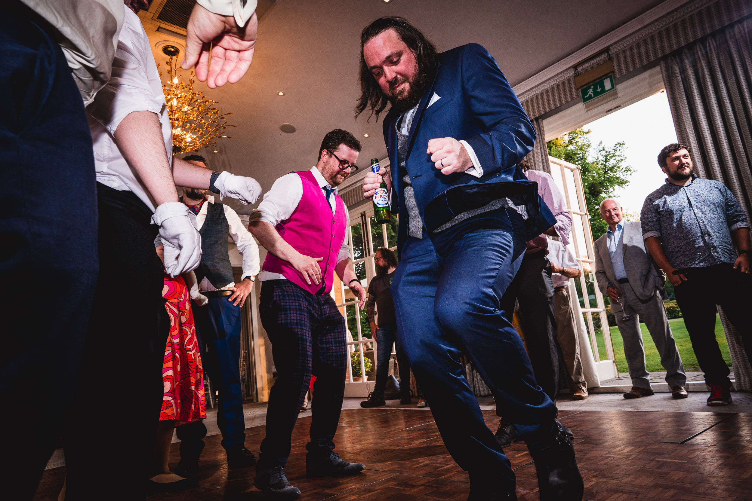 People dancing energetically at an indoor event, with one person wearing a blue suit in the foreground. Other guests and a window with an outdoor view are in the background.