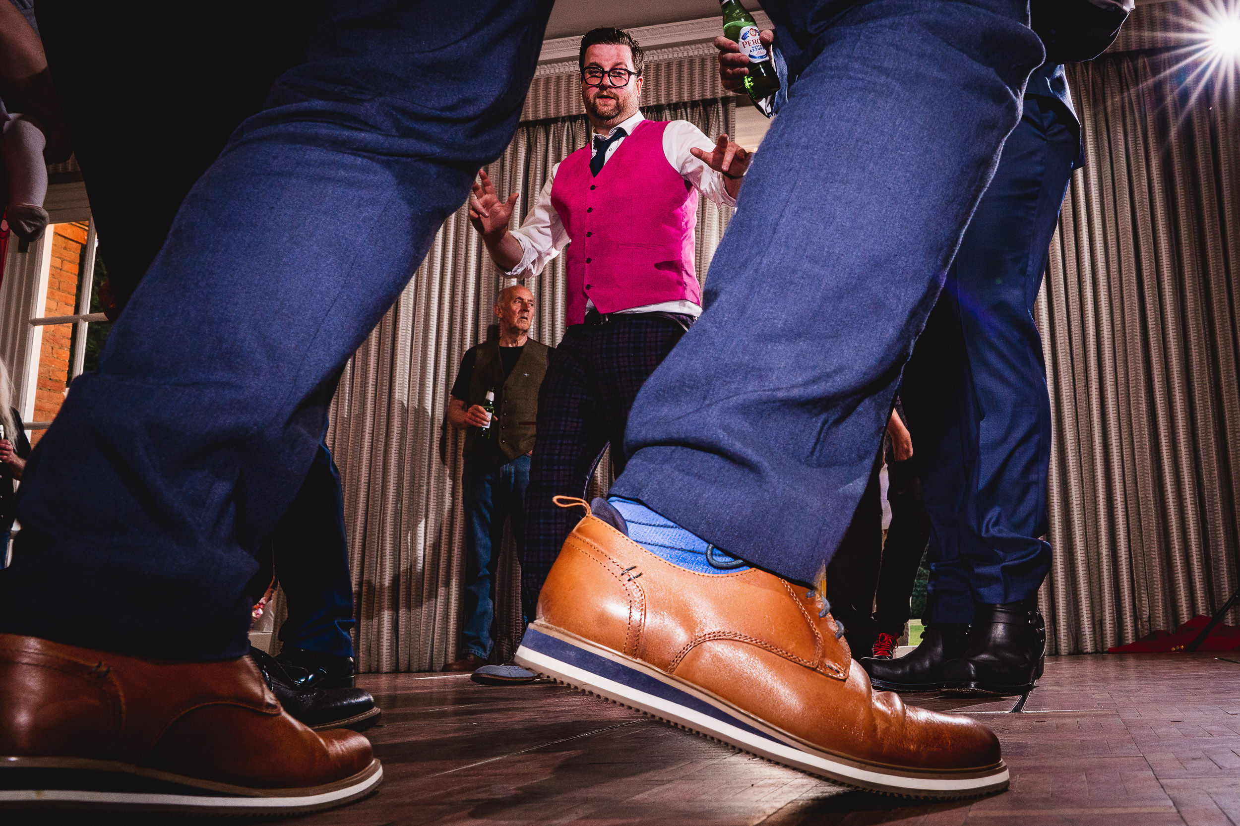 People dancing at an indoor event, with a focus on a person in a bright pink vest and brown shoes.