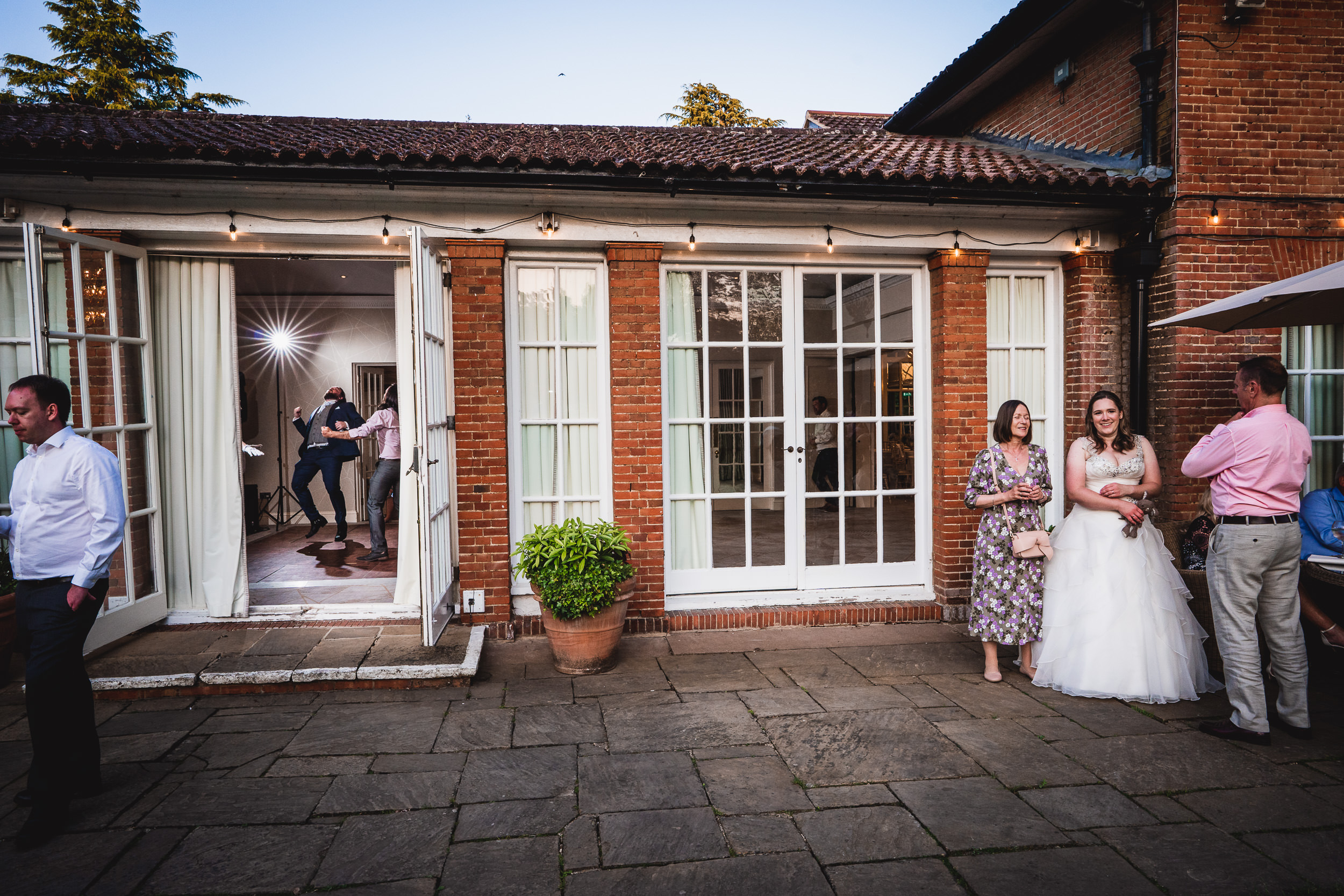 A bride and a few guests stand outside a building with large glass doors, while two people dance inside.