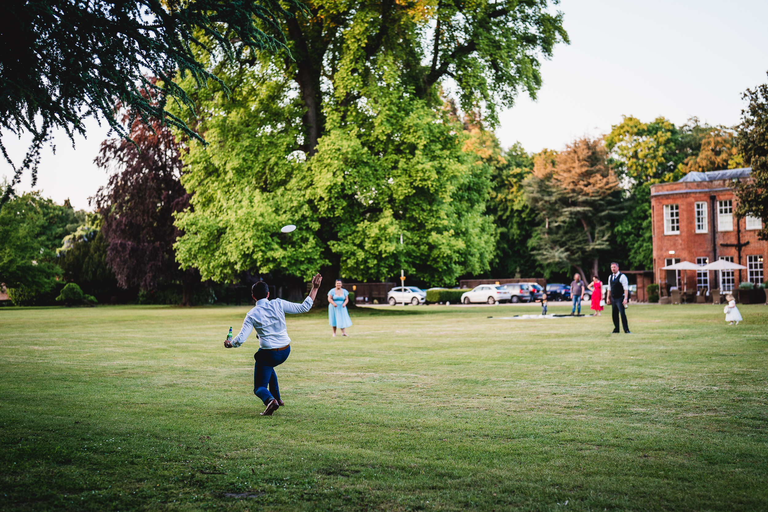 People playing a game of frisbee on a grassy lawn with trees in the background and a brick building to the right.