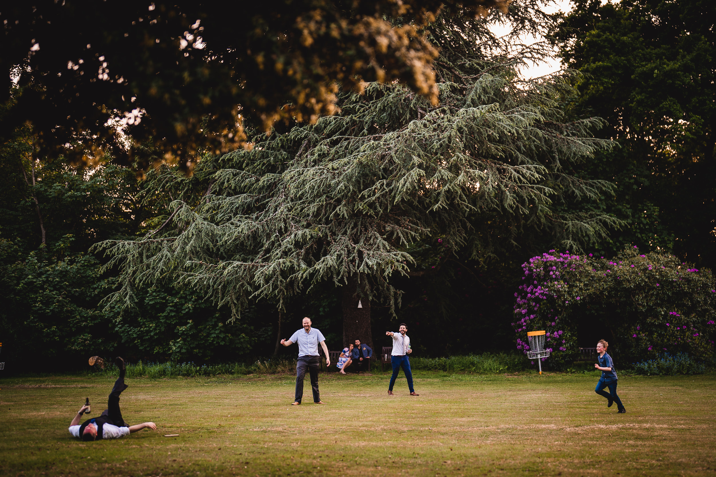 People playing frisbee in a park with a large tree and flowers in the background. One person is on the ground, while others stand nearby.