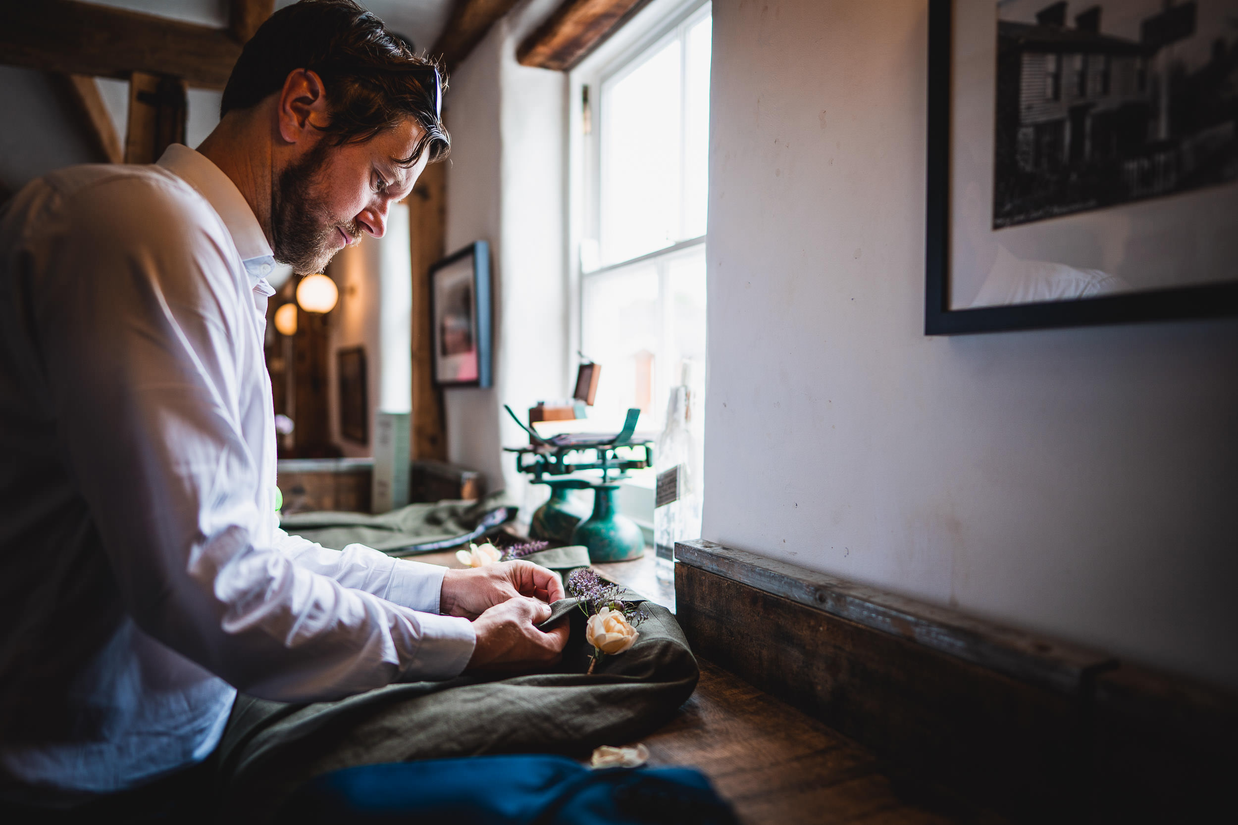 Man in a white shirt works with fabric near a window in a rustic room with wooden beams and framed pictures.