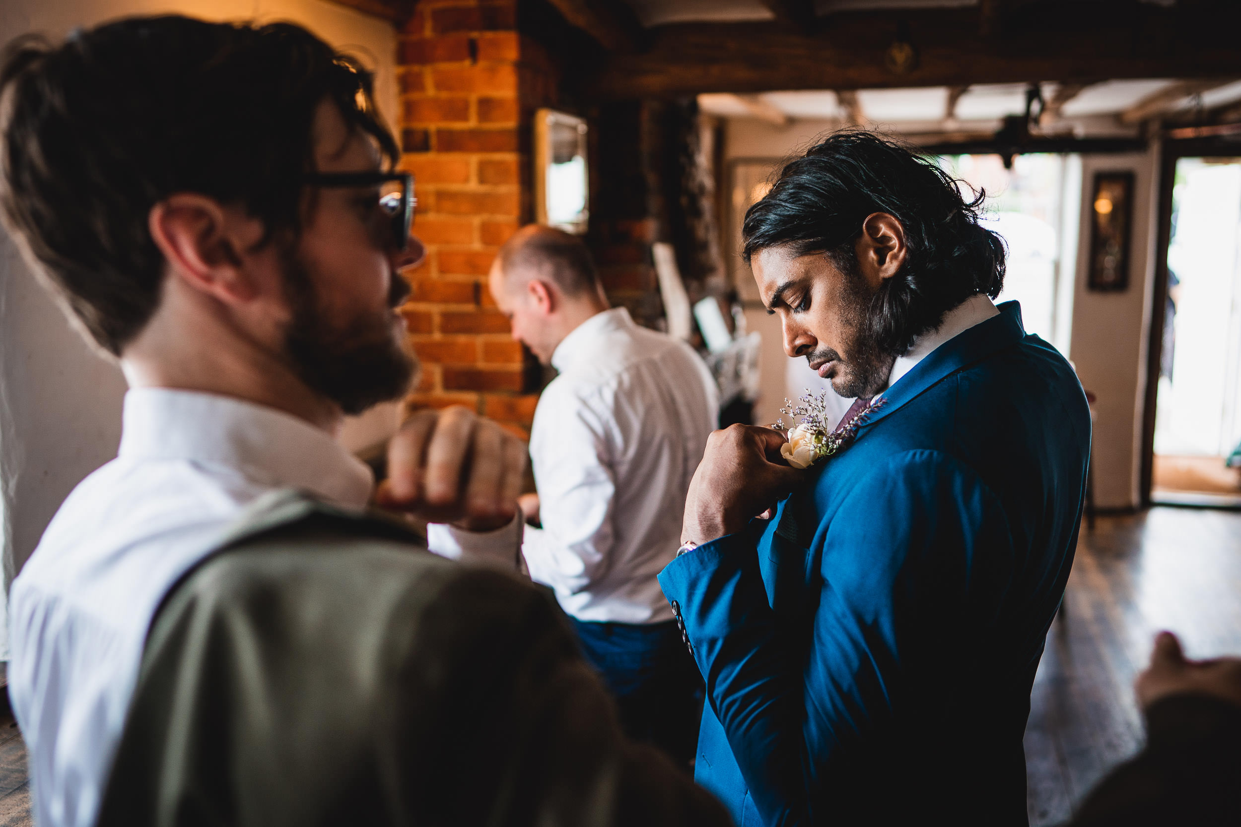 A man in a blue suit adjusts his boutonniere while two other men in white shirts stand nearby in a rustic indoor setting.