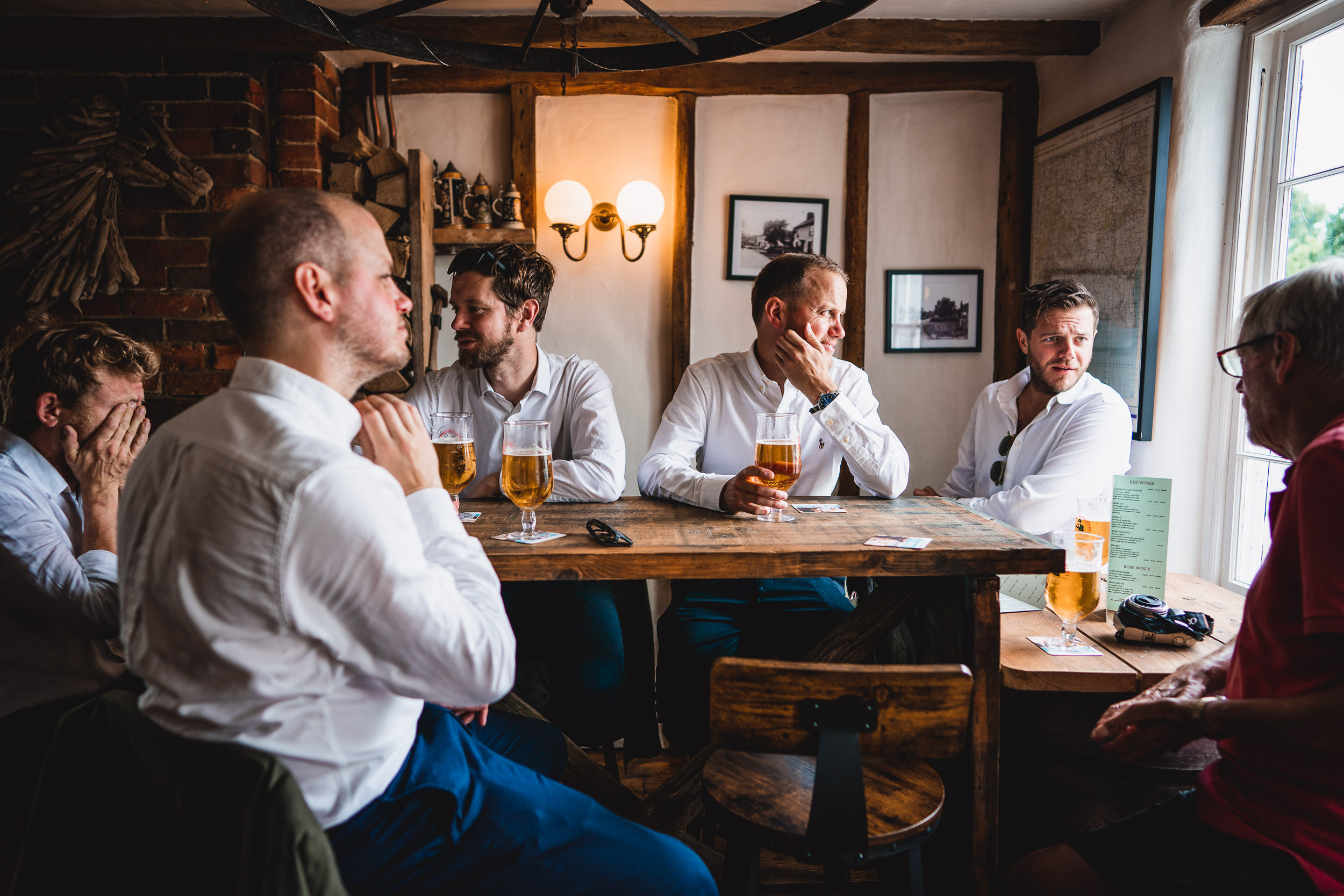 A group of six men sit around a wooden table in a pub, drinking beer and conversing. The room has rustic decor with brick walls and framed pictures. Natural light comes through the window.