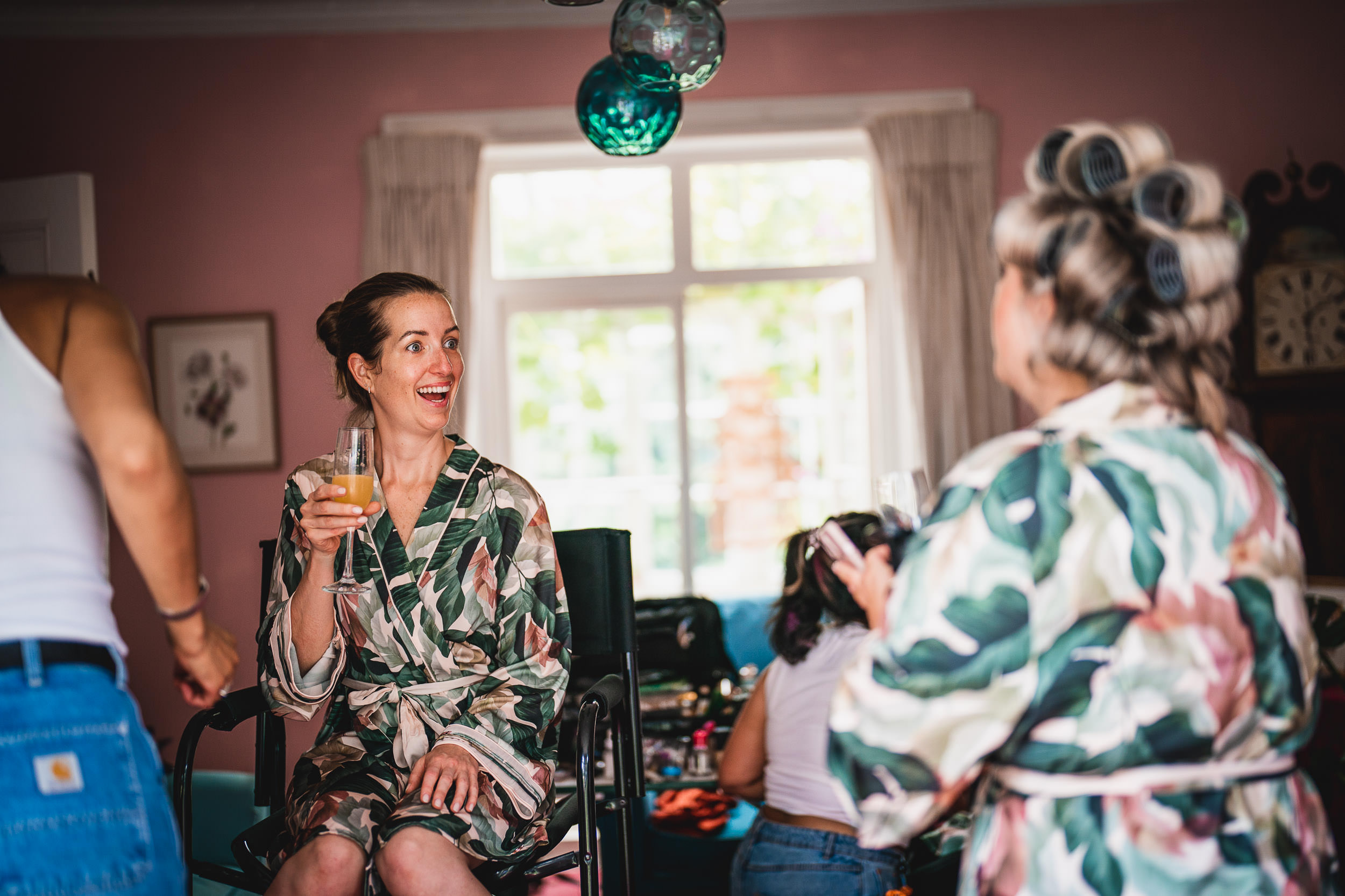 Women in colorful robes with hair rollers chat and smile while holding drinks in a bright room.