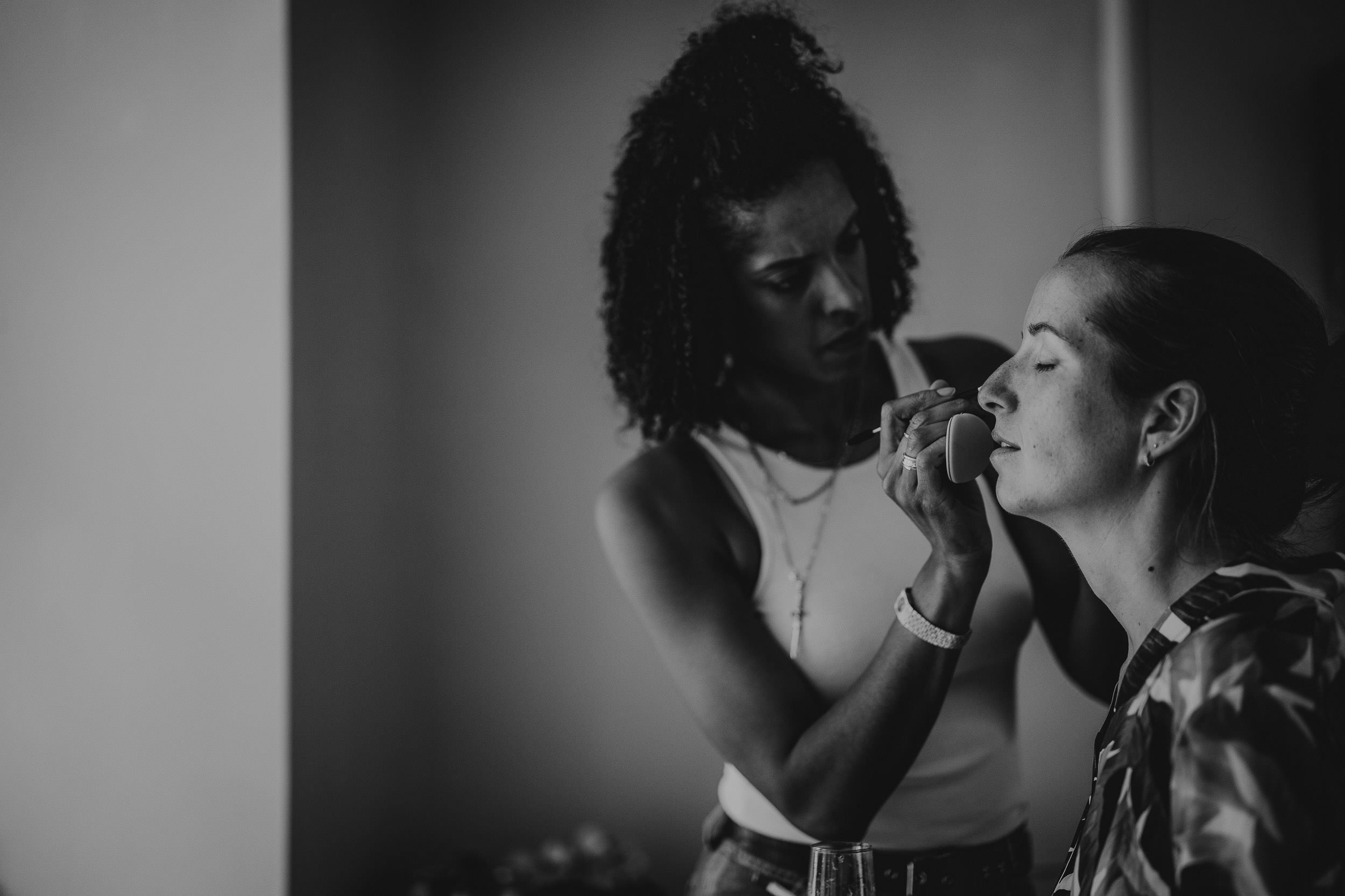 A woman applies makeup to another woman's face using a sponge in a dimly lit room.