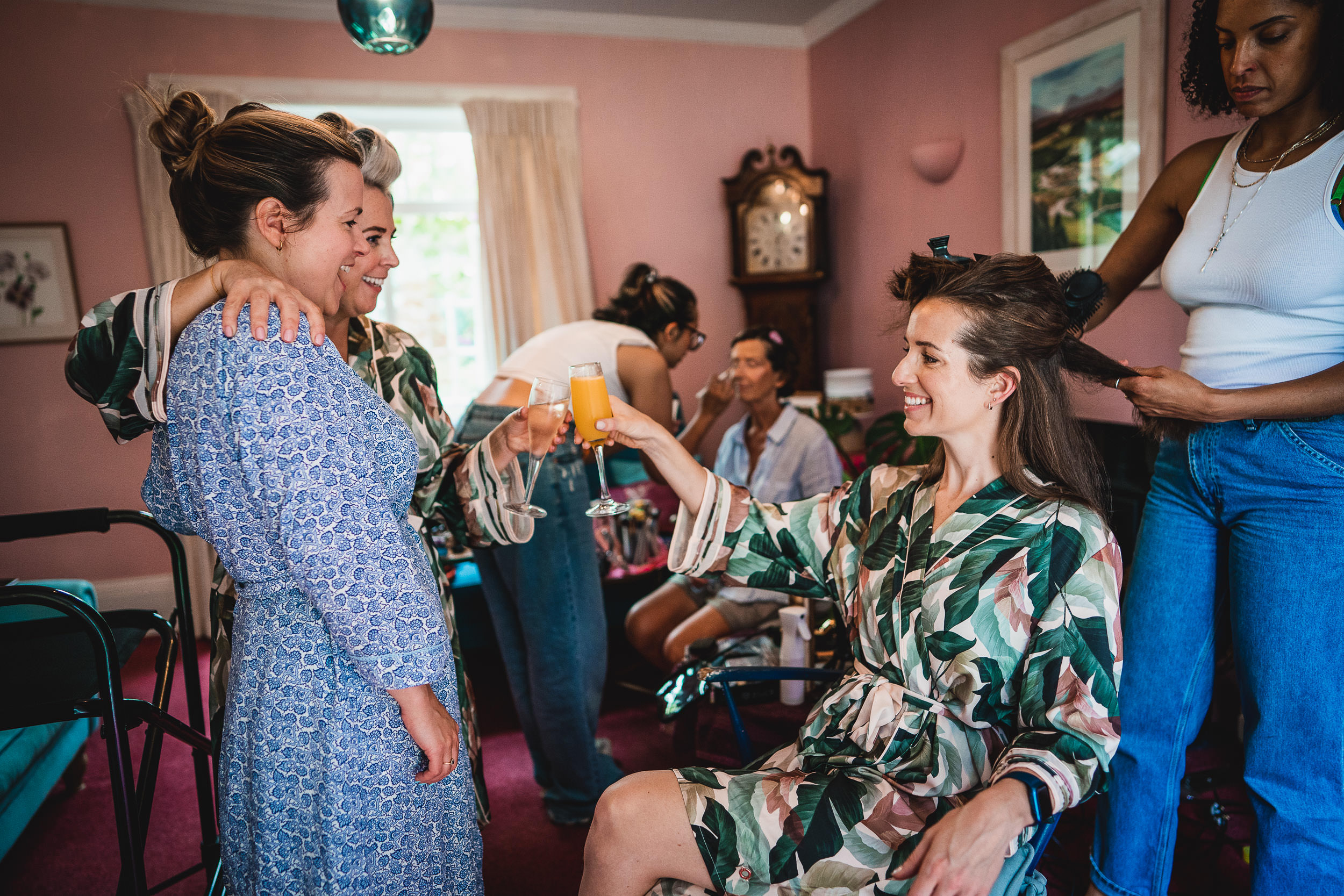 A group of women in robes and casual attire are in a room. One woman holds a drink, while another styles her hair. They all seem to be preparing for an event.