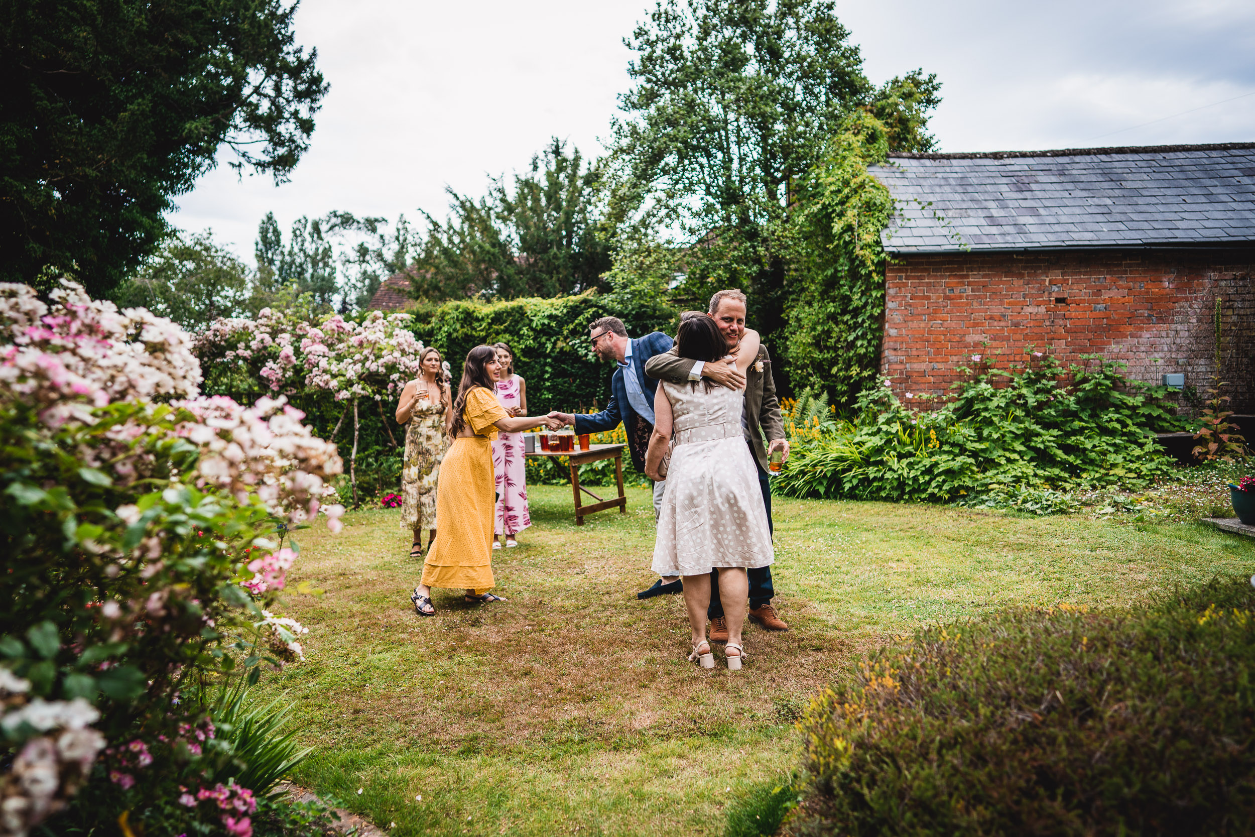 Group of people in a garden greeting and interacting near a table set with drinks. Trees and a brick building form the background.