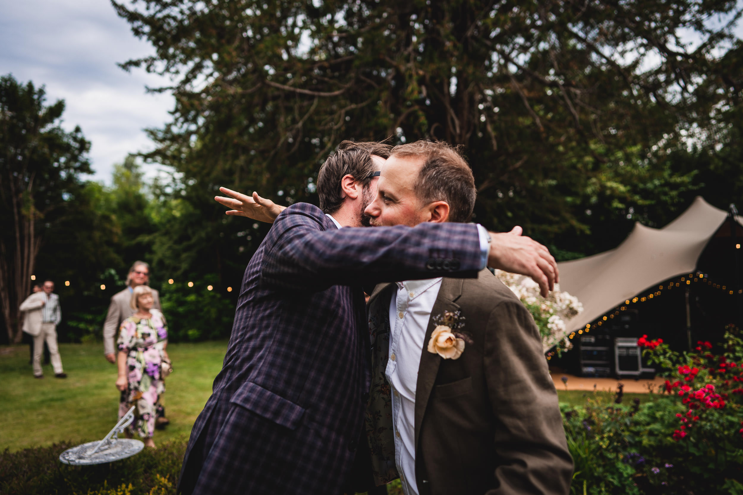 Two men embrace in a hug outdoors, surrounded by trees and a canopy. Other people are visible in the background.