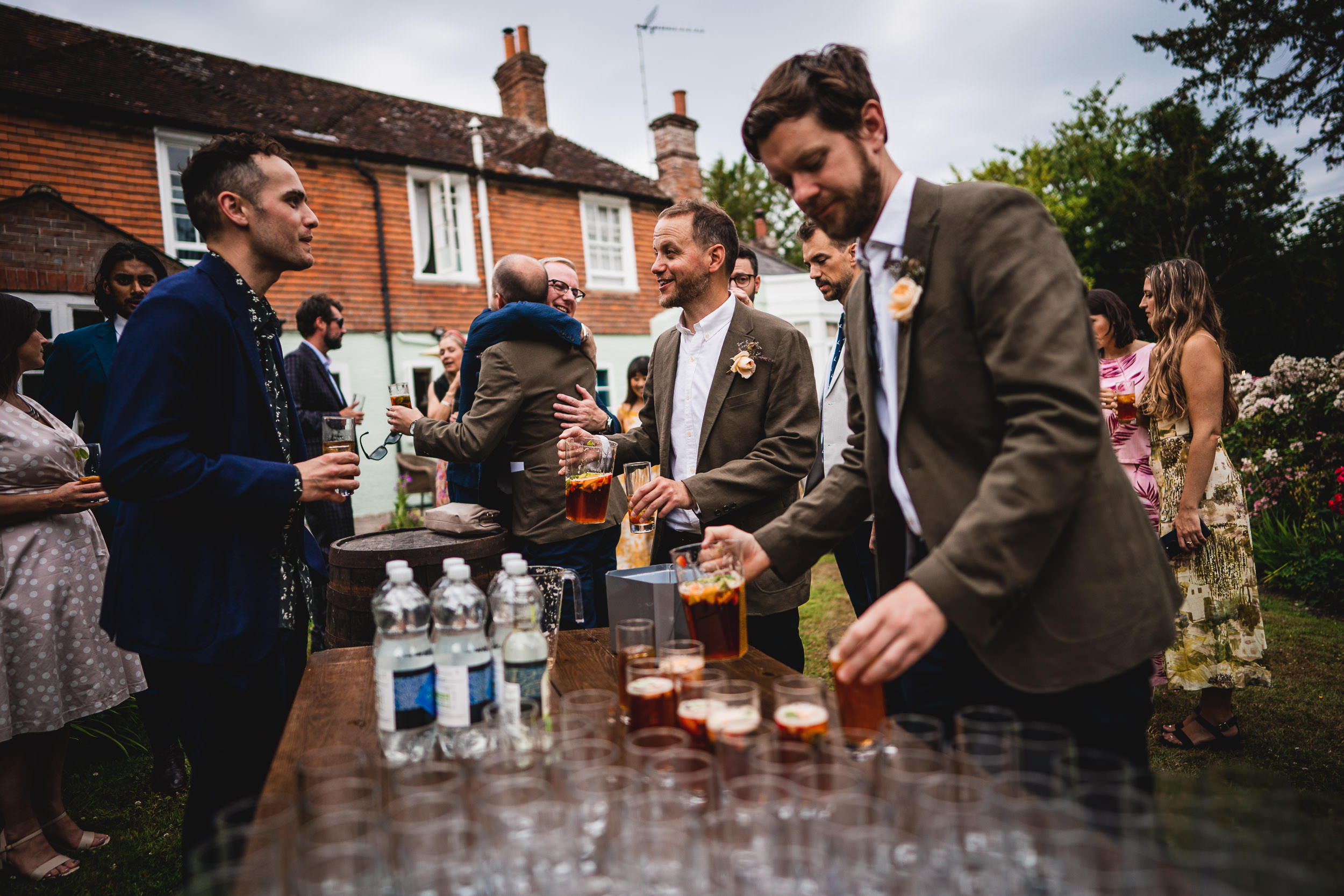 People at an outdoor gathering near a brick house, serving drinks on a wooden table.