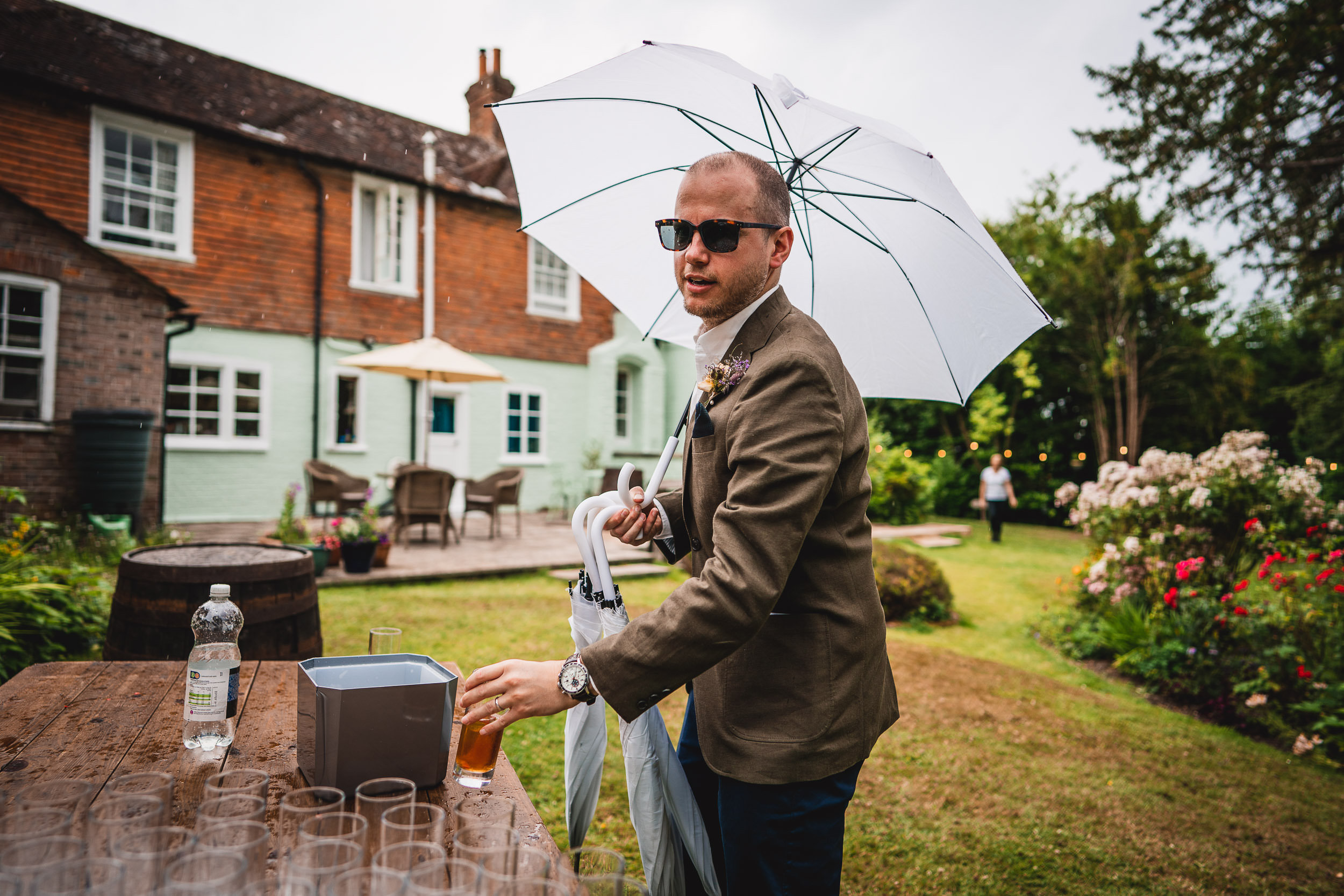 A man in sunglasses holding a white umbrella pours a drink outdoors near a rustic house and garden.