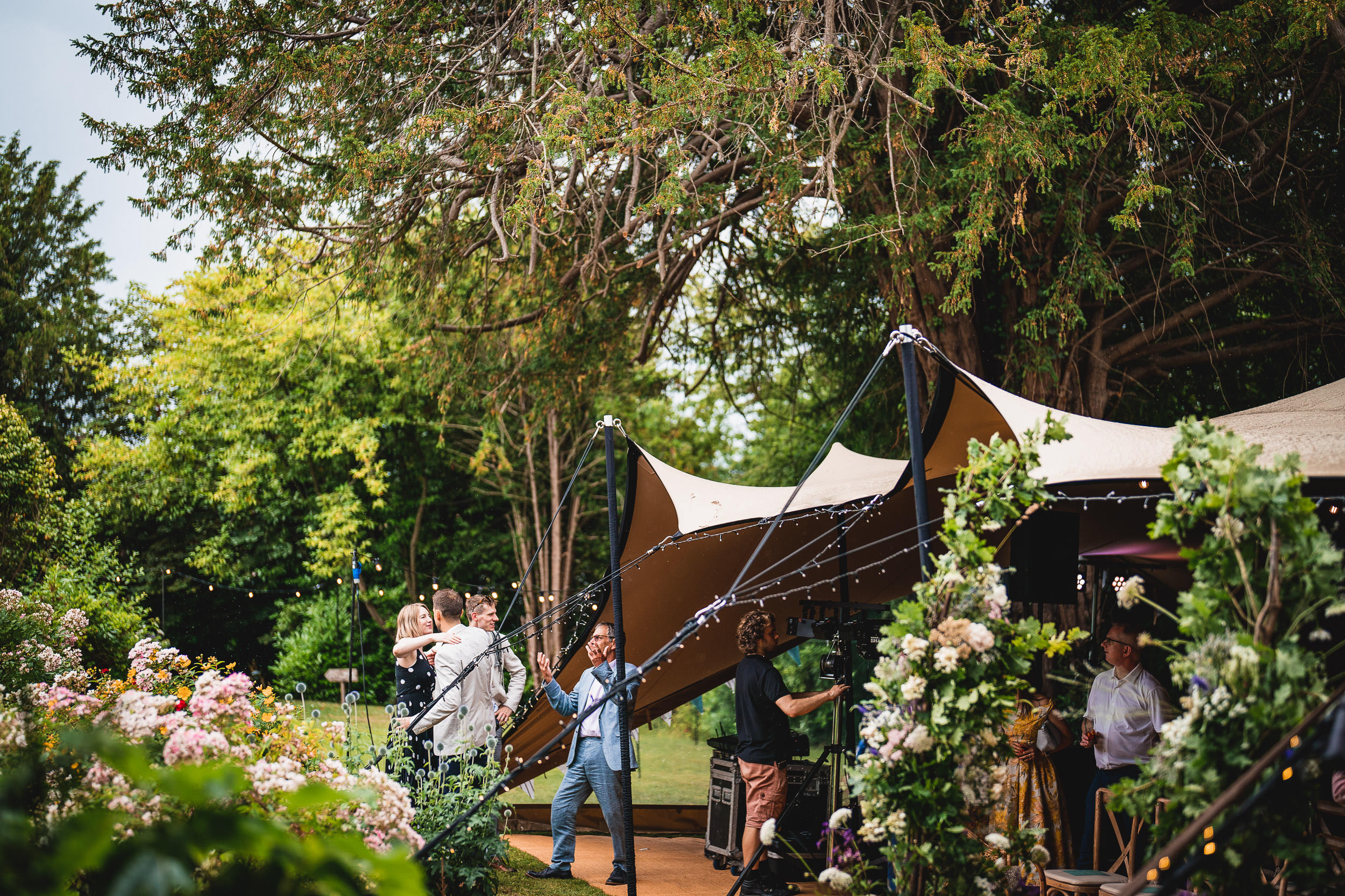 People dancing inside a tent with string lights, surrounded by trees and flowers in a garden setting.