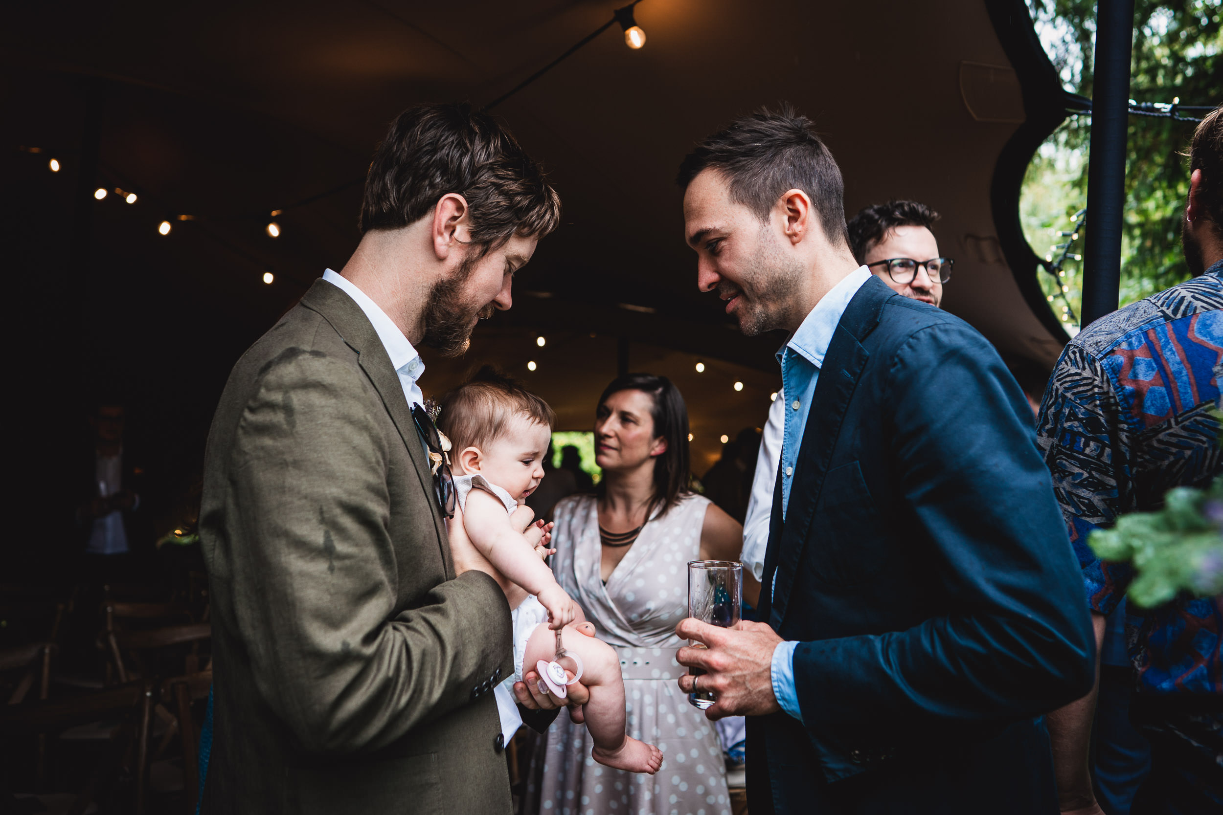 Two men stand conversing at an event, one holding a baby. A woman and another man are in the background, with dim lighting and greenery visible.