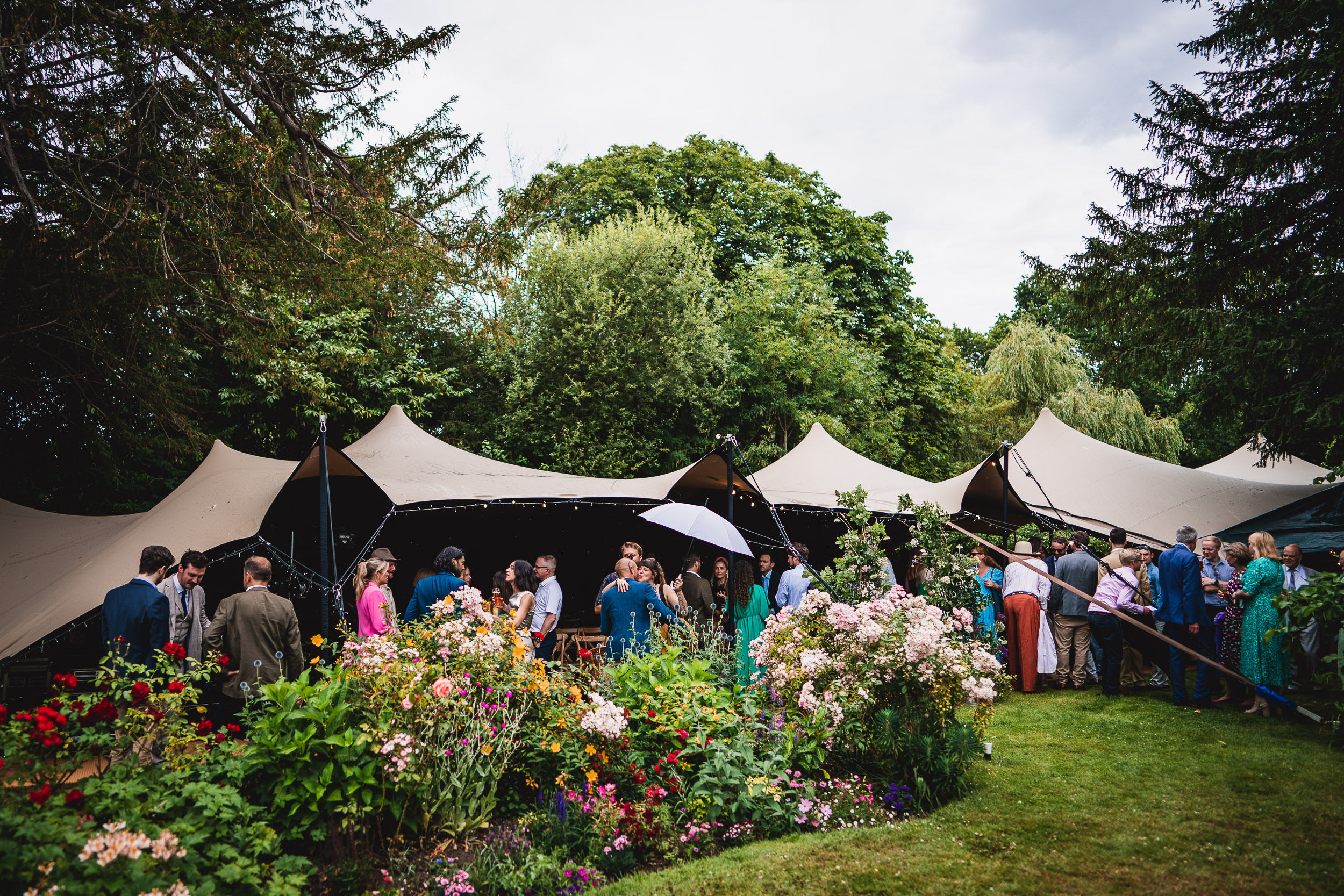 People gather outdoors under large canopy tents surrounded by lush greenery and colorful flowers.