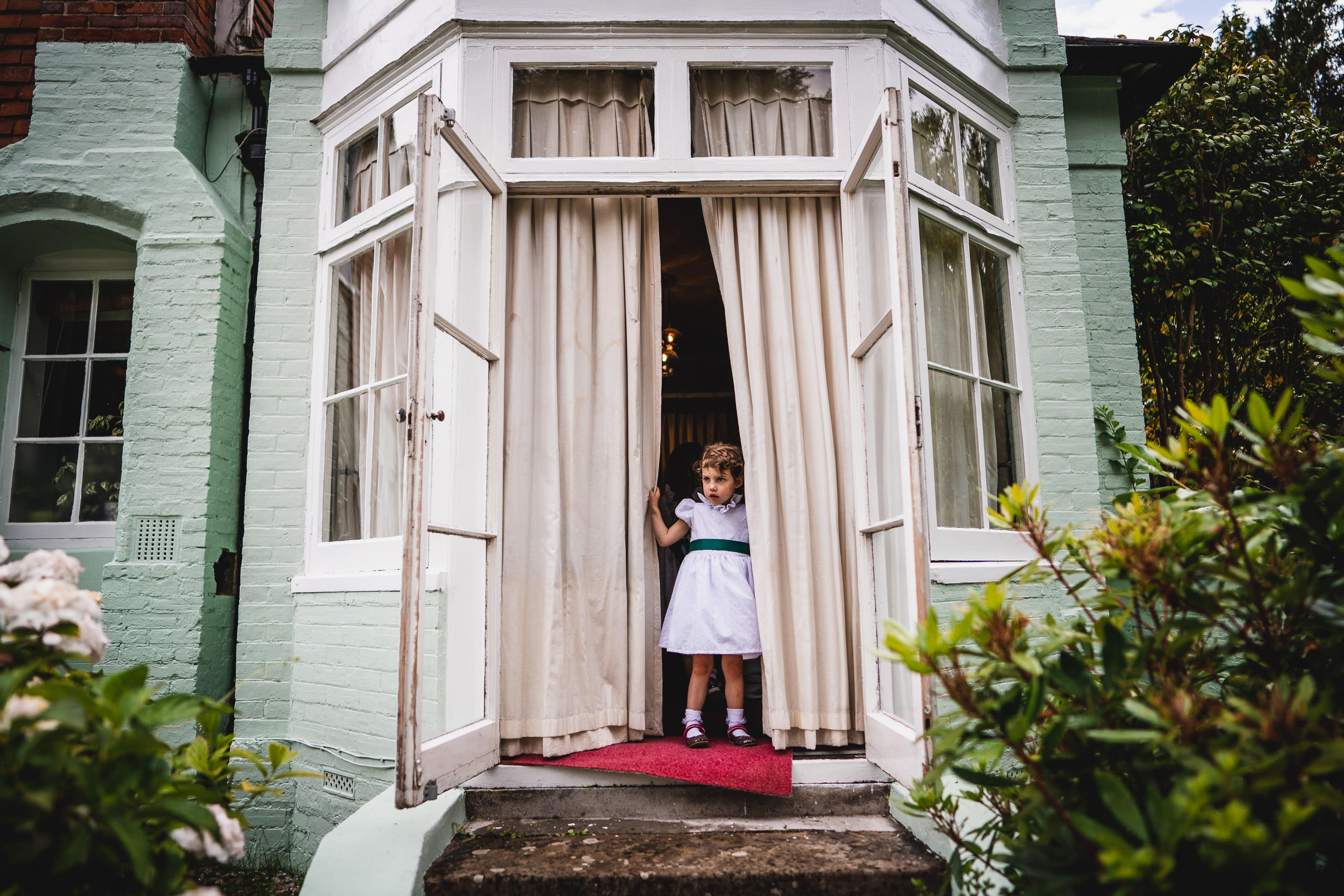 A young girl in a white dress stands in an open doorway with curtains, looking outside. Green plants are in the foreground.