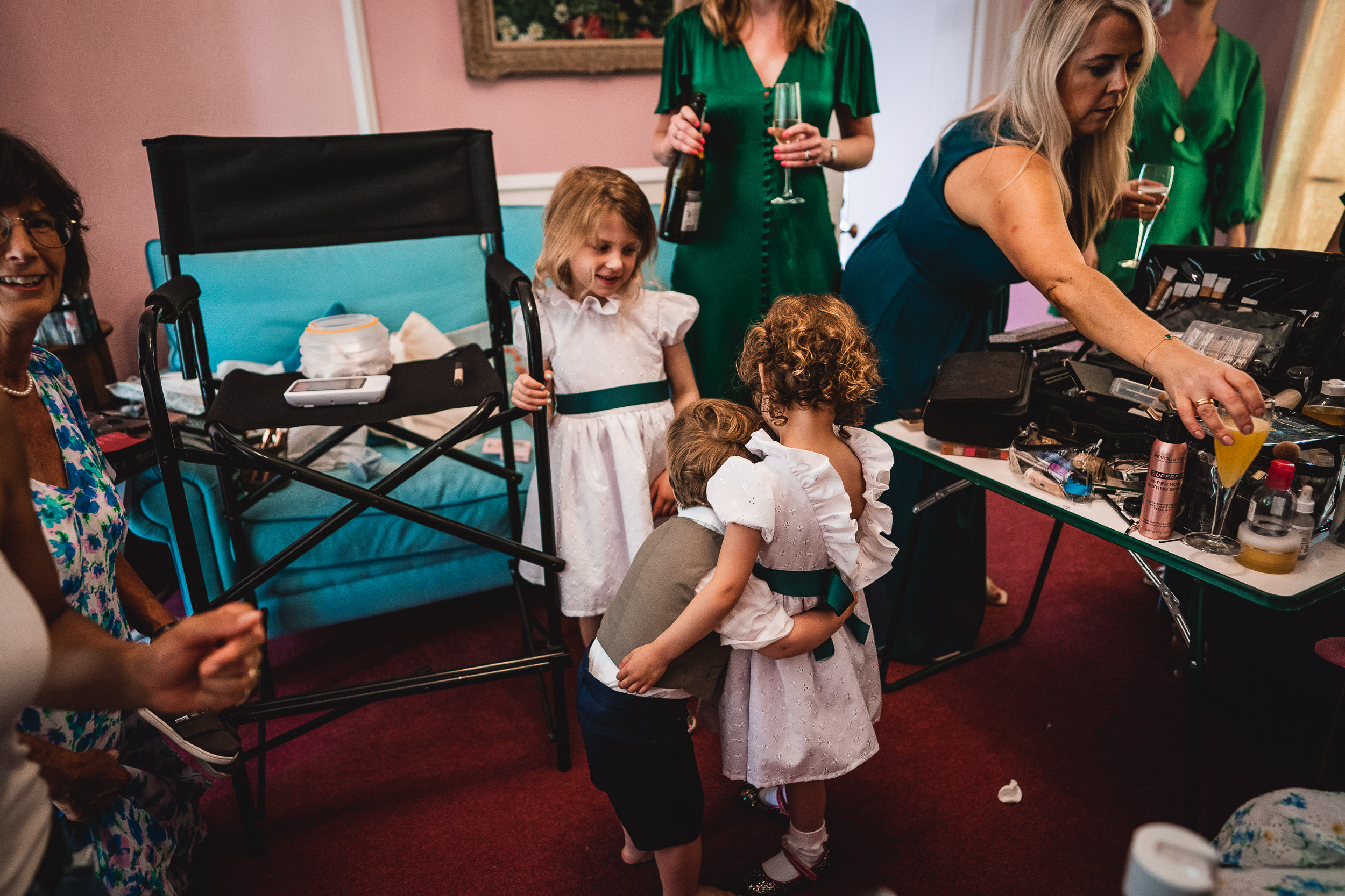 Children embrace in a lively room with adults in dresses, makeup items, and drinks on a table.