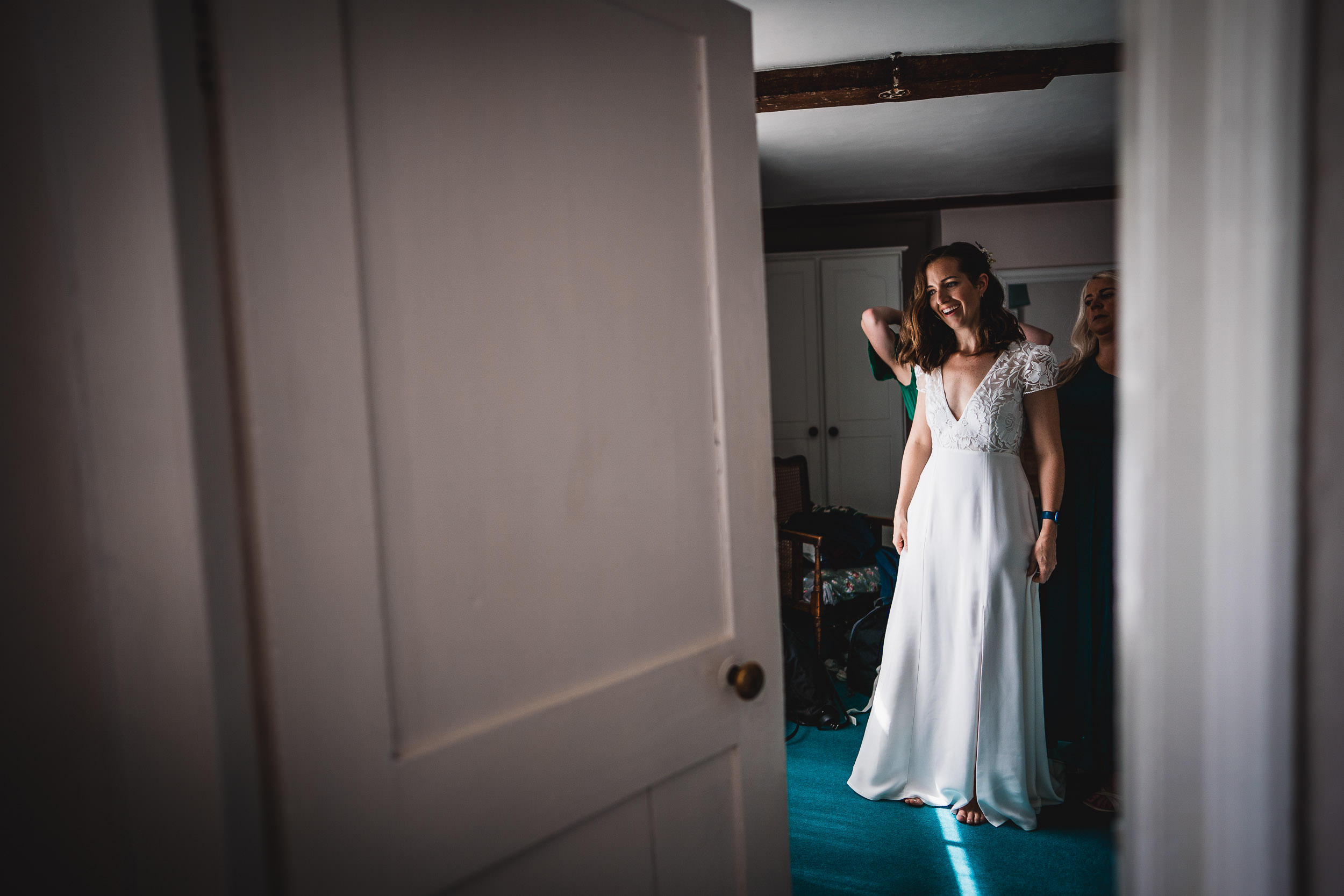 A woman in a white dress stands inside a room with a partially open door, while another person helps with her attire.