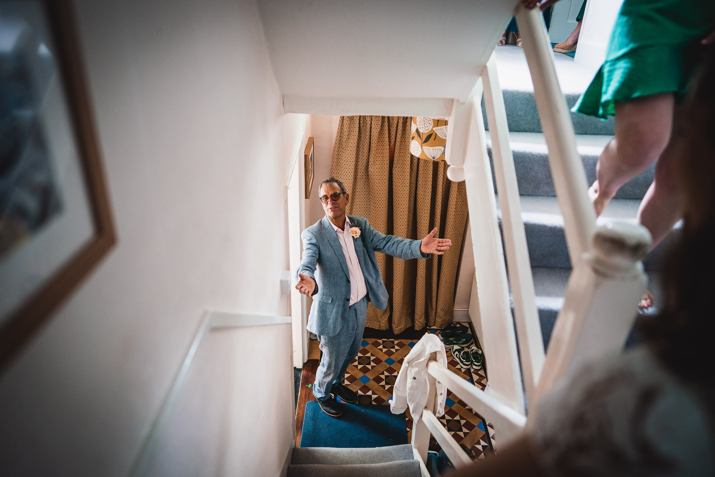 Man in a light blue suit stands at the bottom of a staircase with arms open, surrounded by people on stairs.