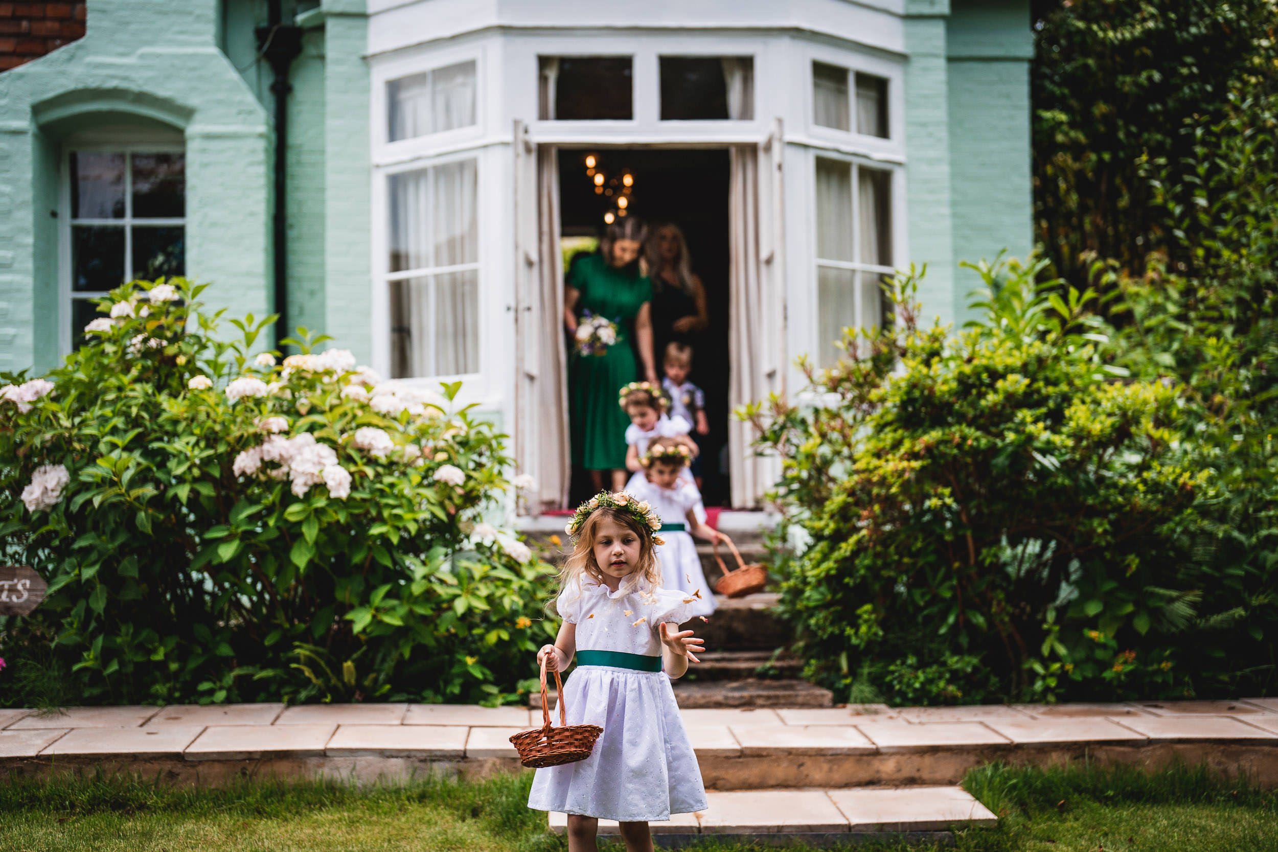 Children in white dresses walk out of a house carrying small baskets, with lush greenery and a brick pathway in the foreground.