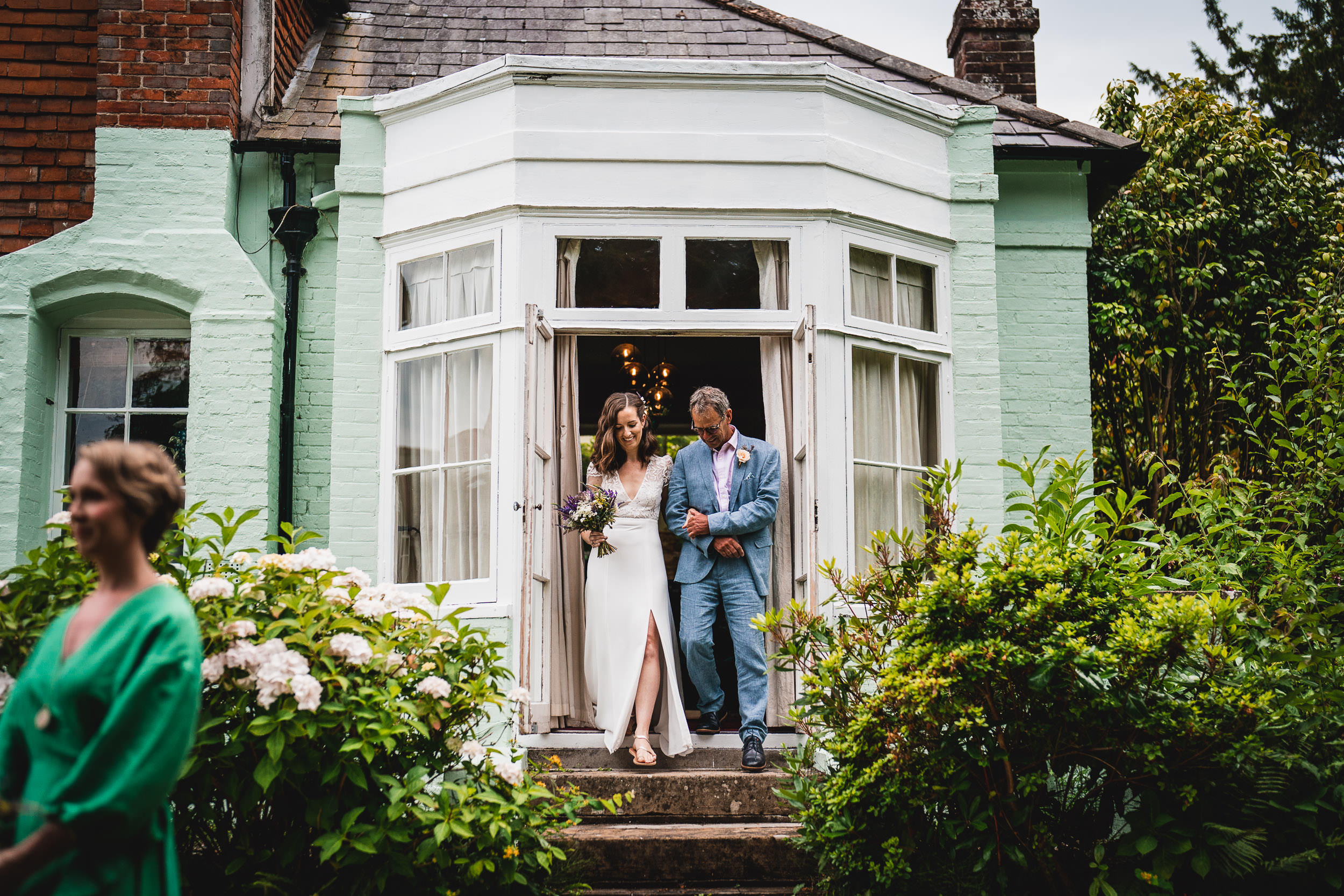 A bride and a man walk out of a house with a green wall, surrounded by greenery and flowers. A woman in a green dress stands in the foreground.