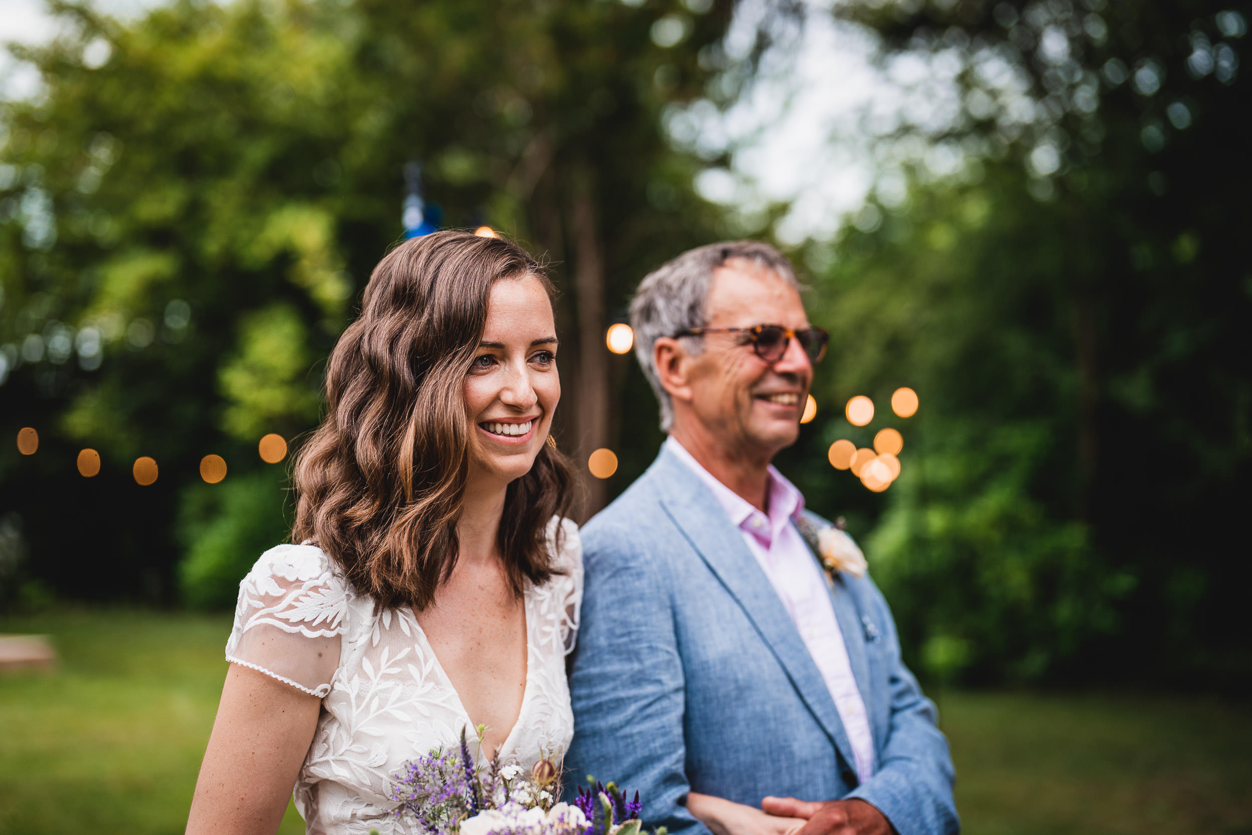 A woman in a white dress and man in sunglasses smile outdoors, with string lights and greenery in the background.