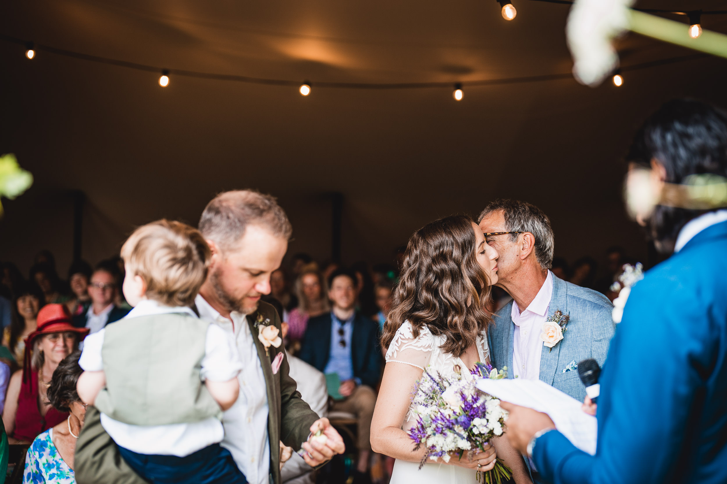 A bride and groom share a kiss during a wedding ceremony. A man holding a child stands nearby. Guests are seated under a tent adorned with string lights.