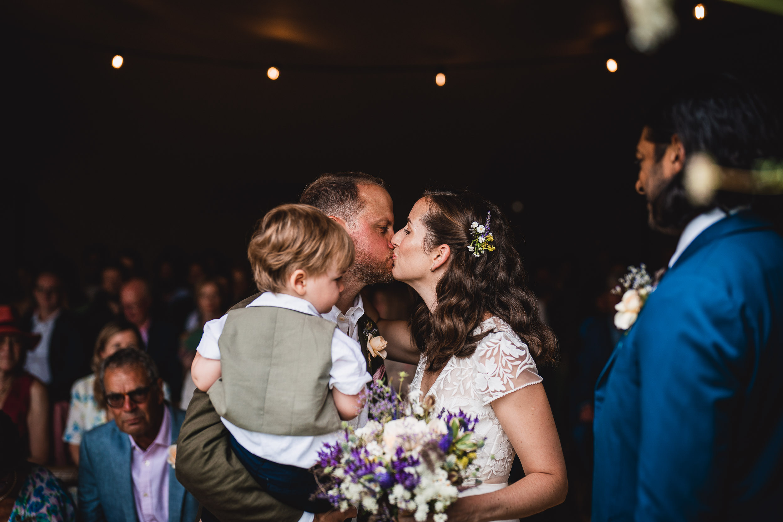 A couple kisses at their wedding ceremony, holding a child. The groom is in a suit and the bride wears a white dress, holding flowers. Guests are seated and watching.