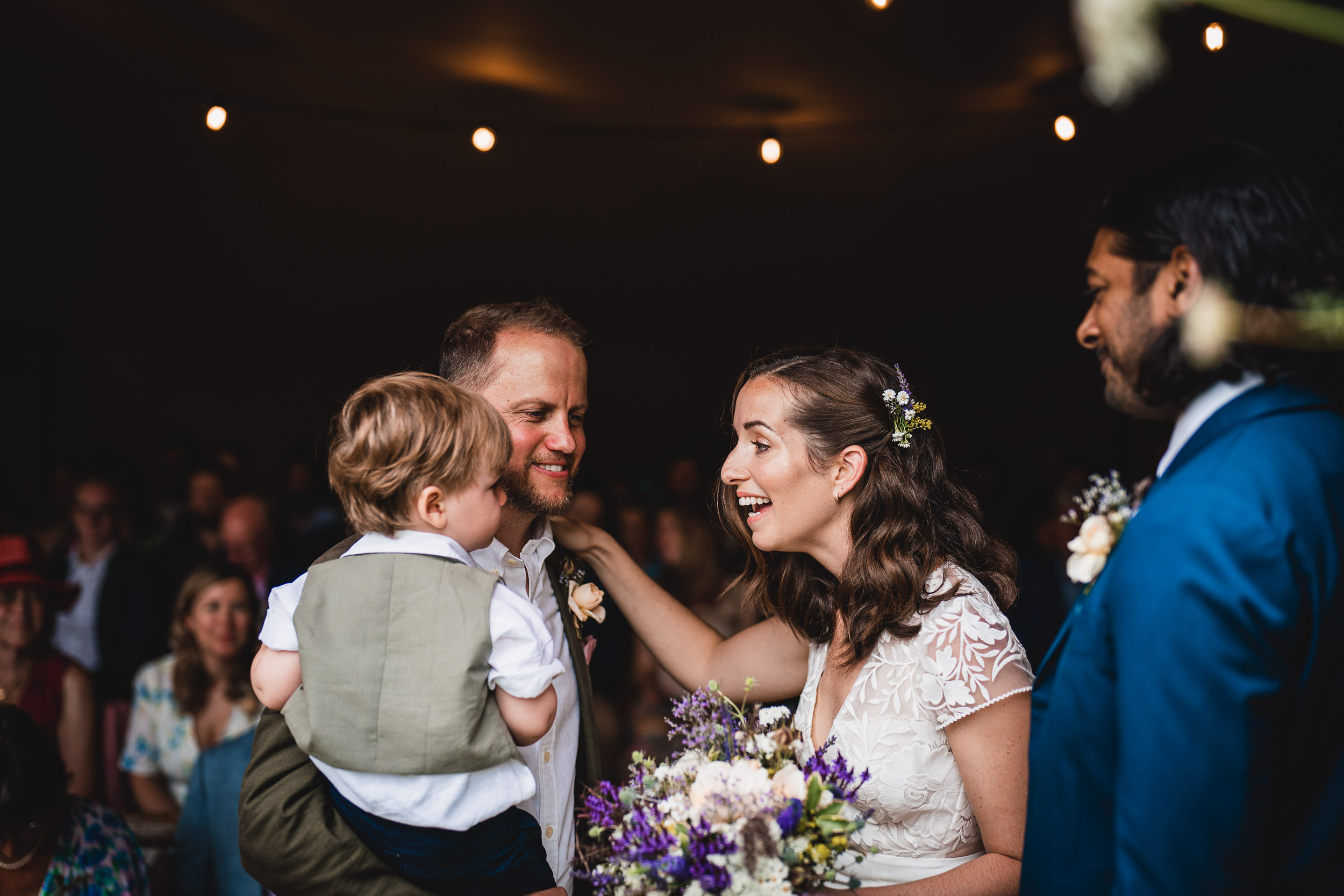 Bride and groom share a joyful moment at their wedding, surrounded by guests. The groom holds a child as the bride smiles, holding a bouquet of flowers.