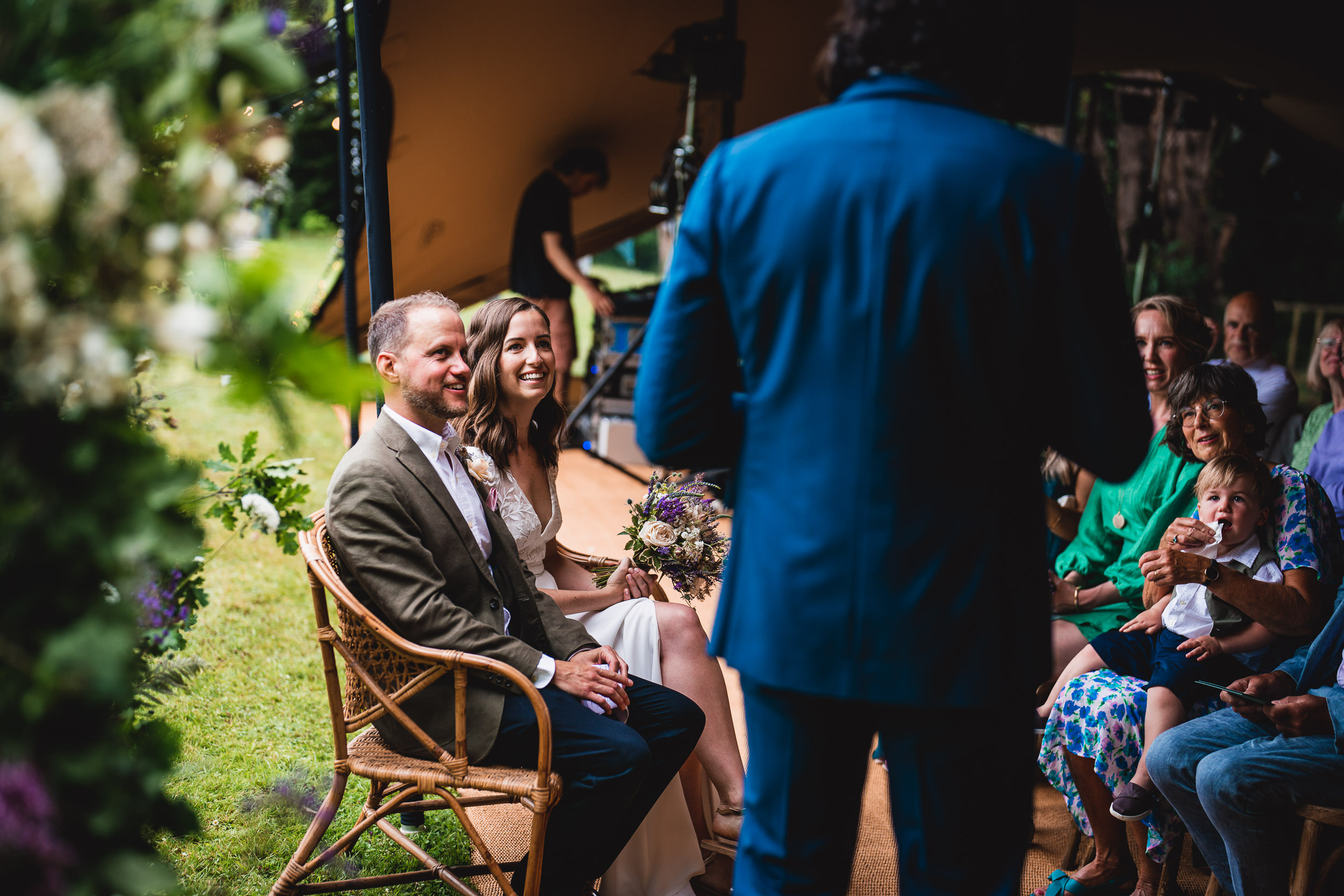 A couple sits at their wedding ceremony, surrounded by guests. A person in a blue suit stands in front of them, likely speaking or officiating. They are seated outdoors.