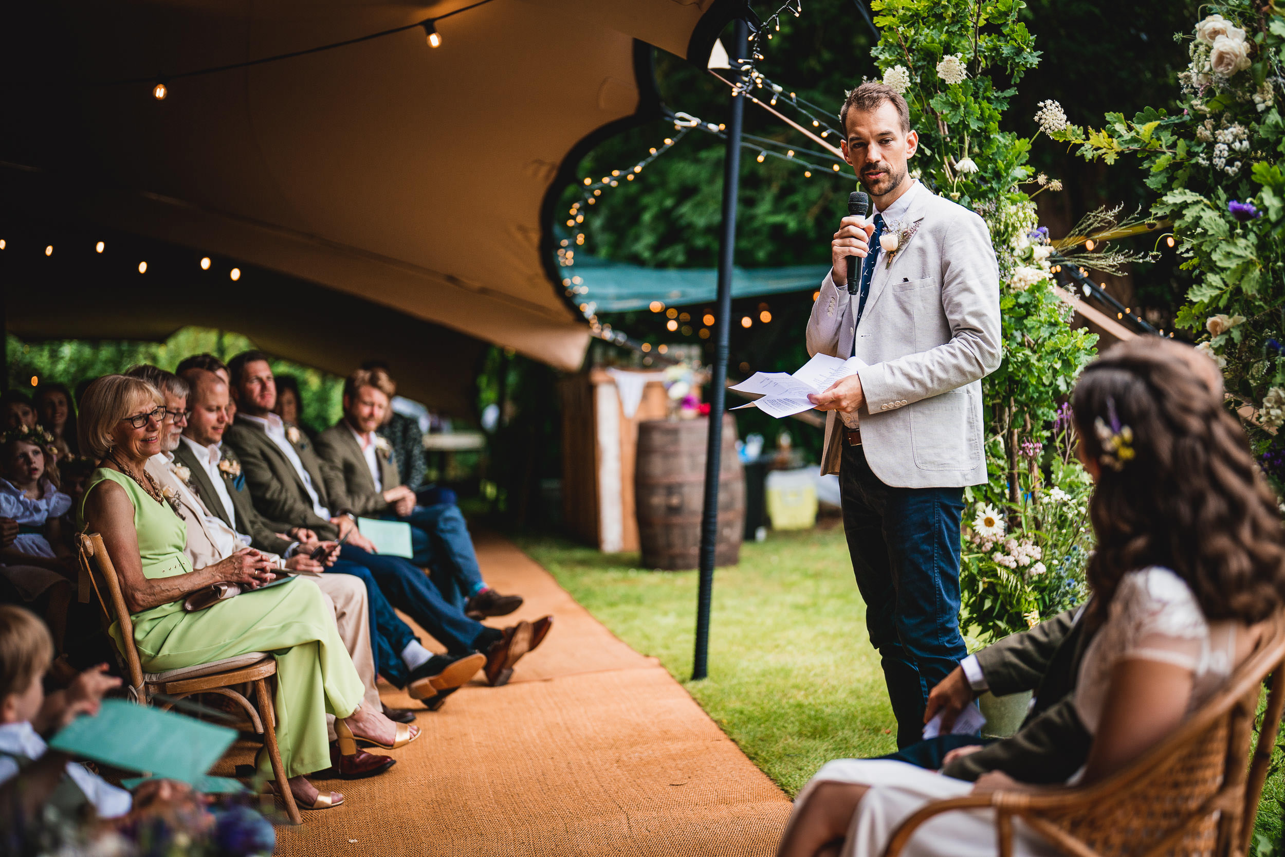 A person stands with a microphone, delivering a speech in a decorated outdoor setting with seated guests.