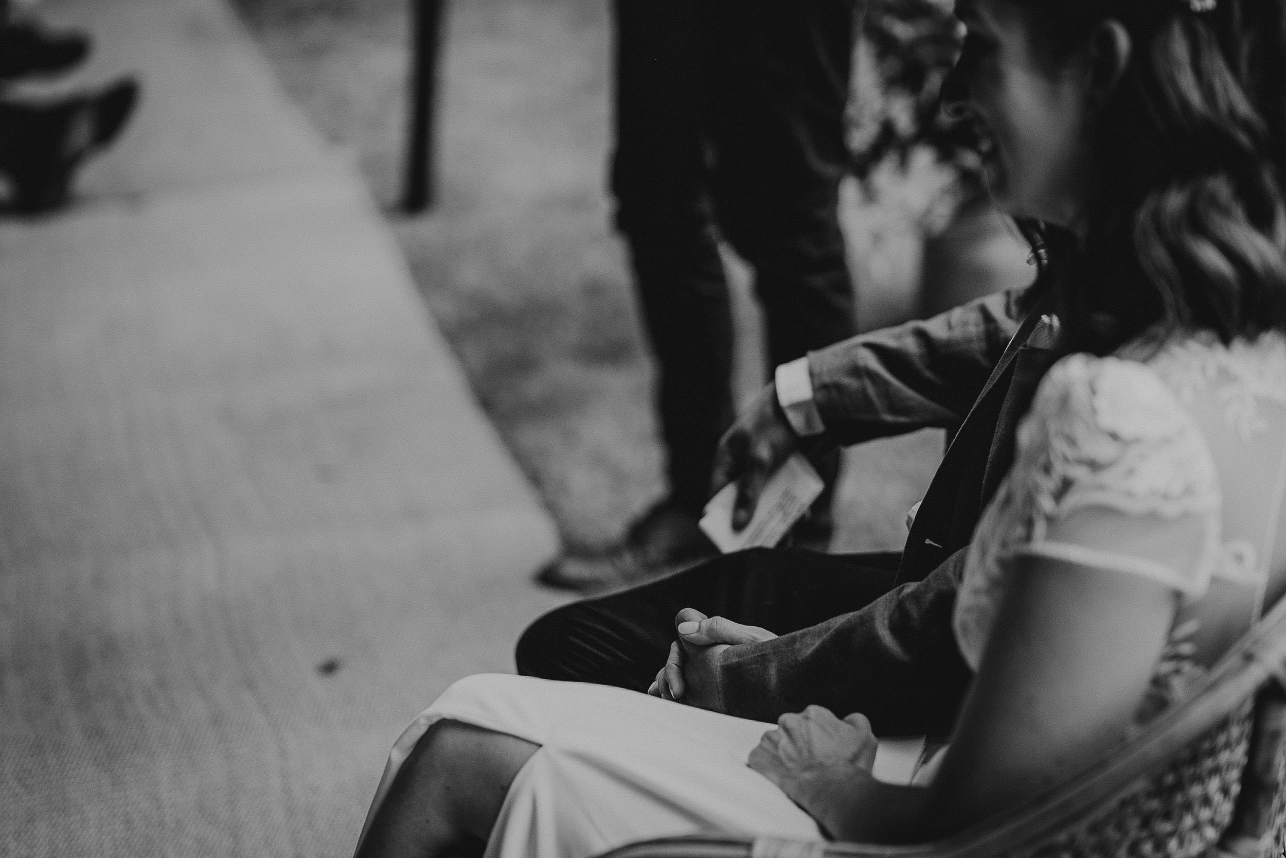 A black and white photo of a couple seated side by side, holding hands, partially visible. They appear to be at a formal event.