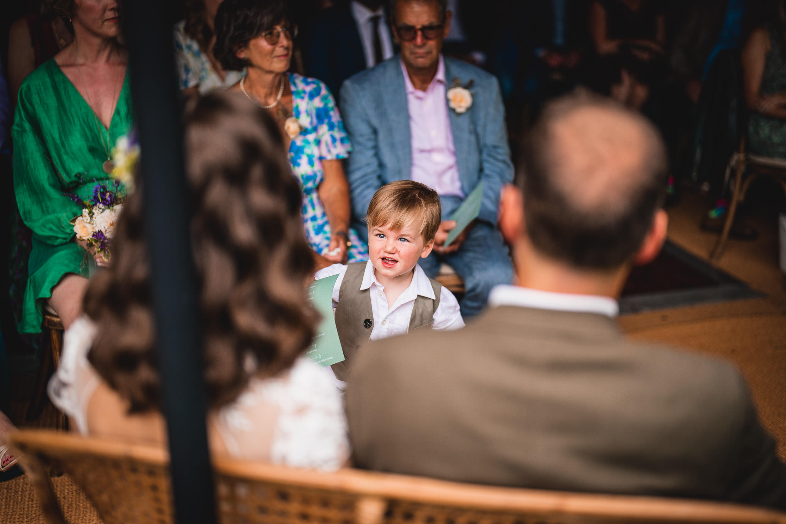 A young child in a vest stands among seated adults during an indoor gathering.