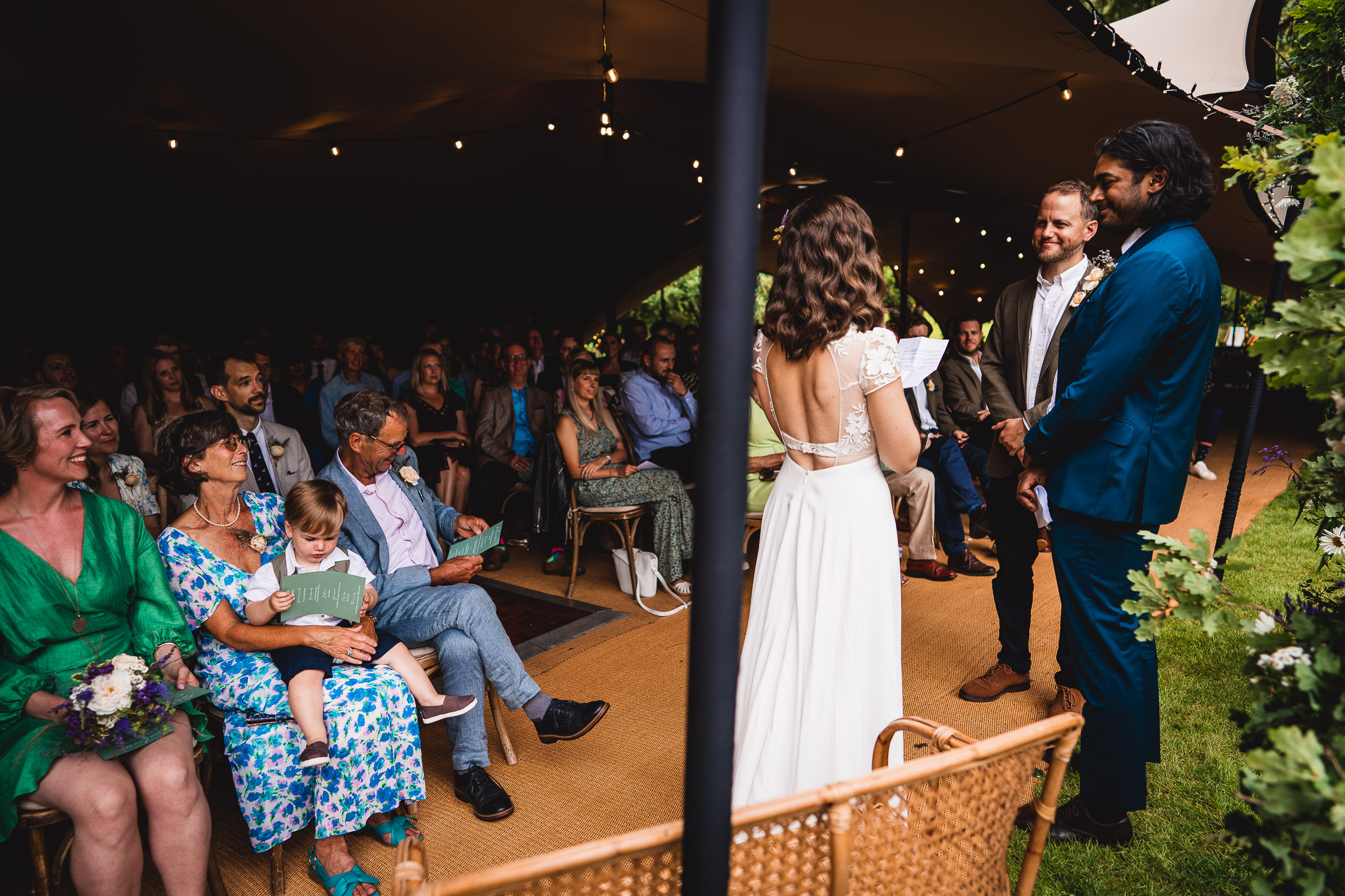 A couple stands facing each other under a tent during a wedding ceremony, with seated guests watching.