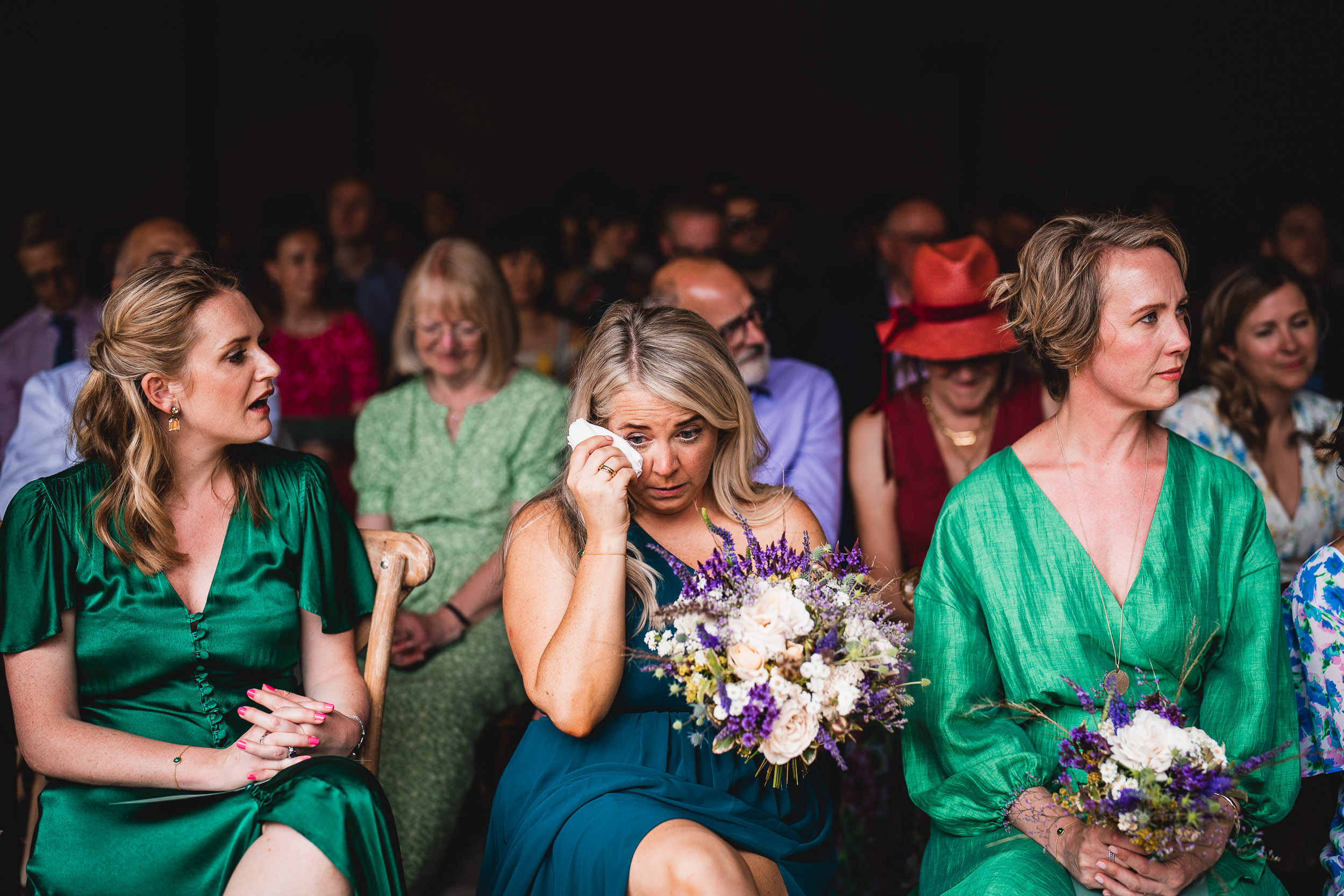 A woman in a green dress wipes a tear while holding a bouquet, seated among people. Two women sit on either side in similar attire, with a group of people visible in the background.