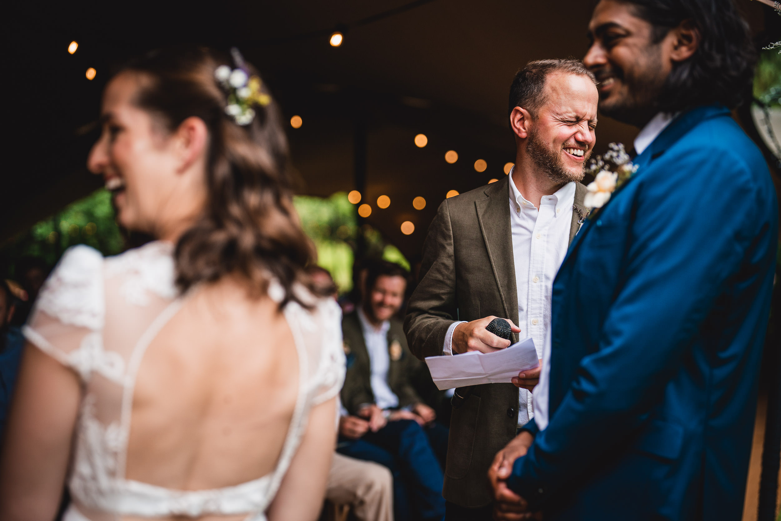 People are attending an outdoor wedding ceremony. A man in a green jacket is holding papers and smiling, while standing near a couple facing each other.